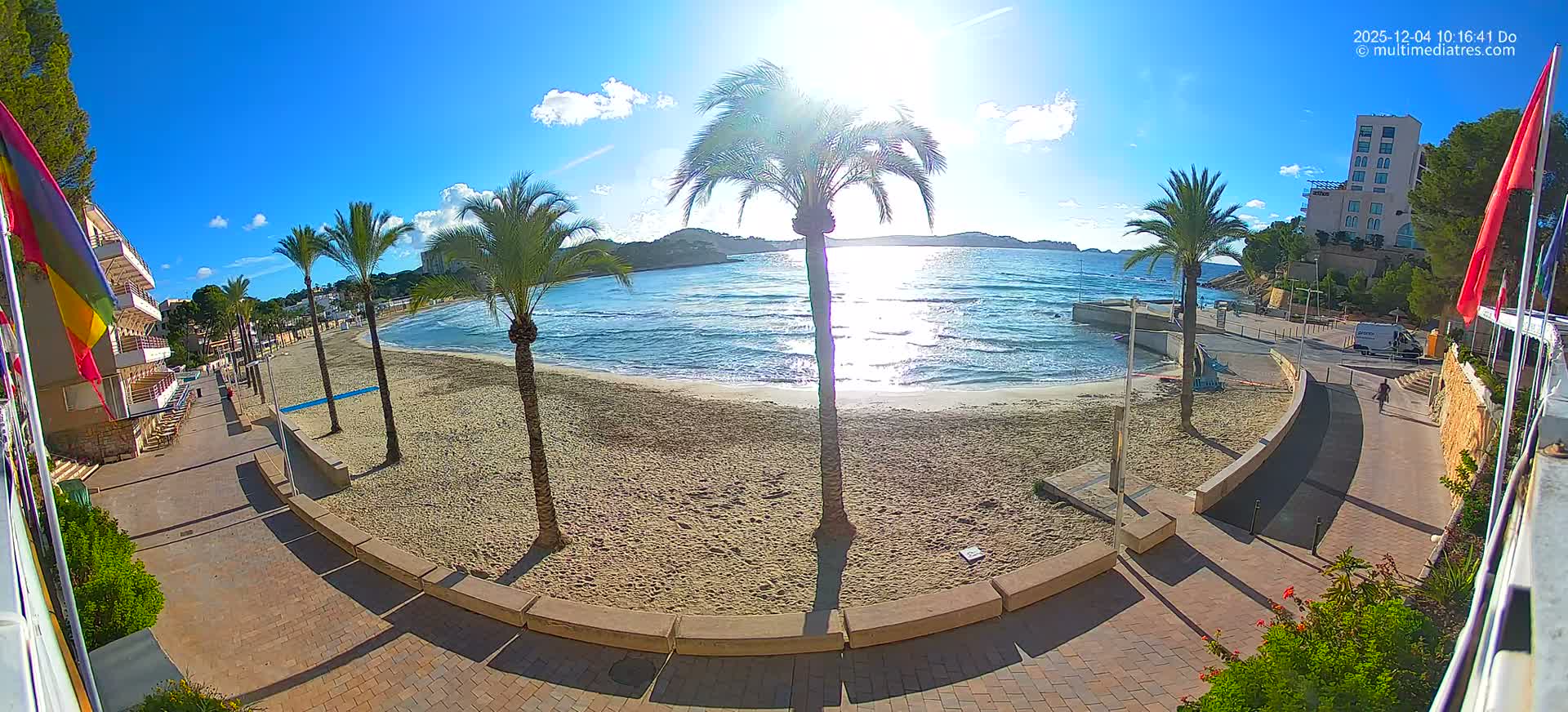 A bright, sunny day with clear blue skies and scattered clouds illuminates a wide sandy beach lined with palm trees, gentle ocean waves, and coastal buildings along a curving promenade.
