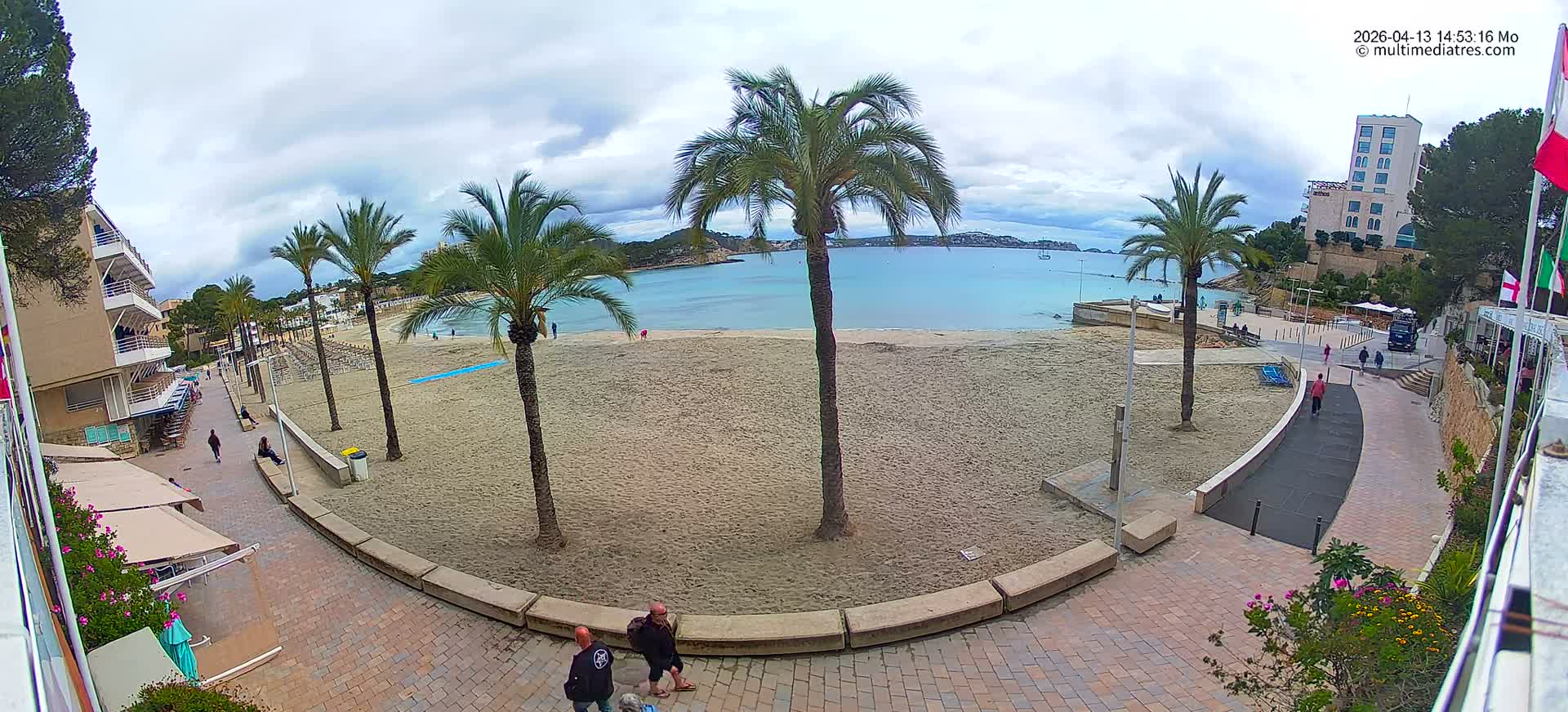 A bright, sunny day with clear blue skies and scattered clouds illuminates a wide sandy beach lined with palm trees, gentle ocean waves, and coastal buildings along a curving promenade.