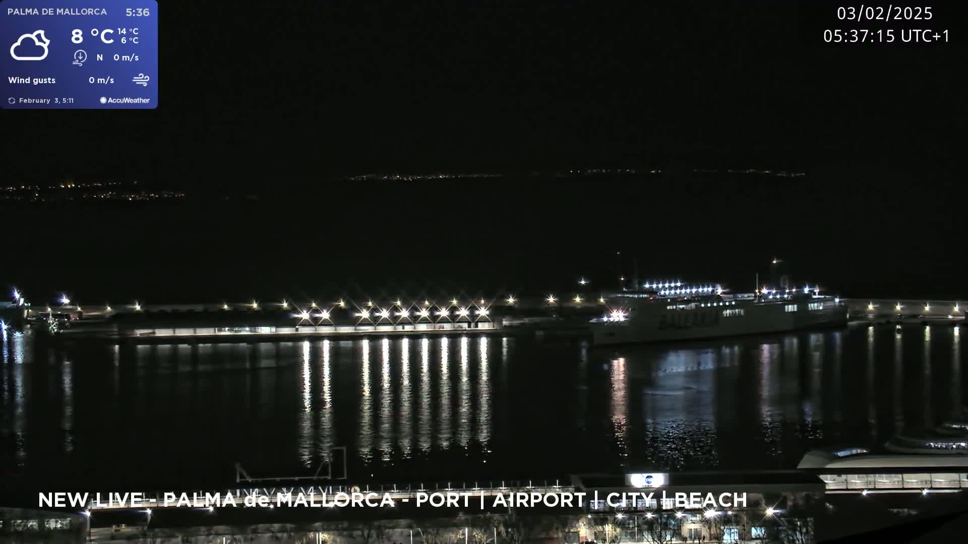 A nighttime view of a harbor with a large ship docked, calm water reflecting lights from the dock and the distant city.