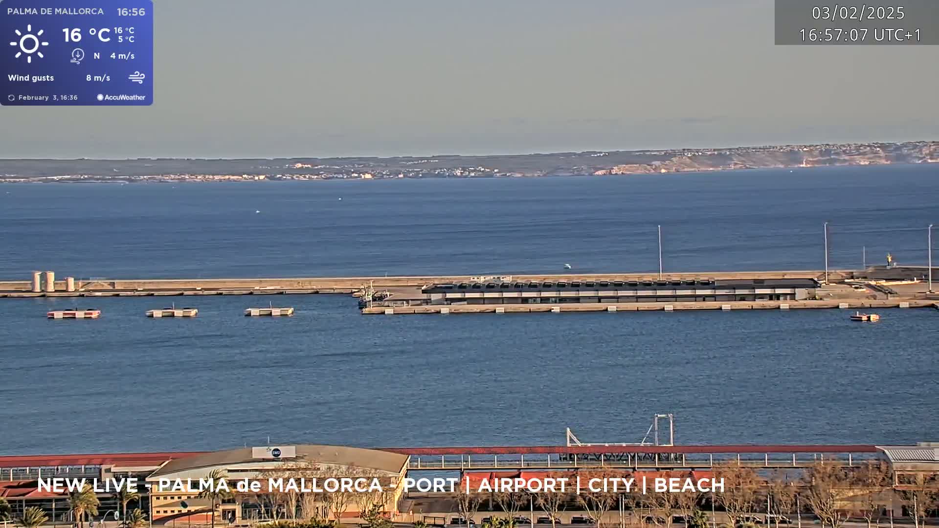 A harbor scene on a sunny day shows calm water, a long pier with several structures, and a distant shoreline.