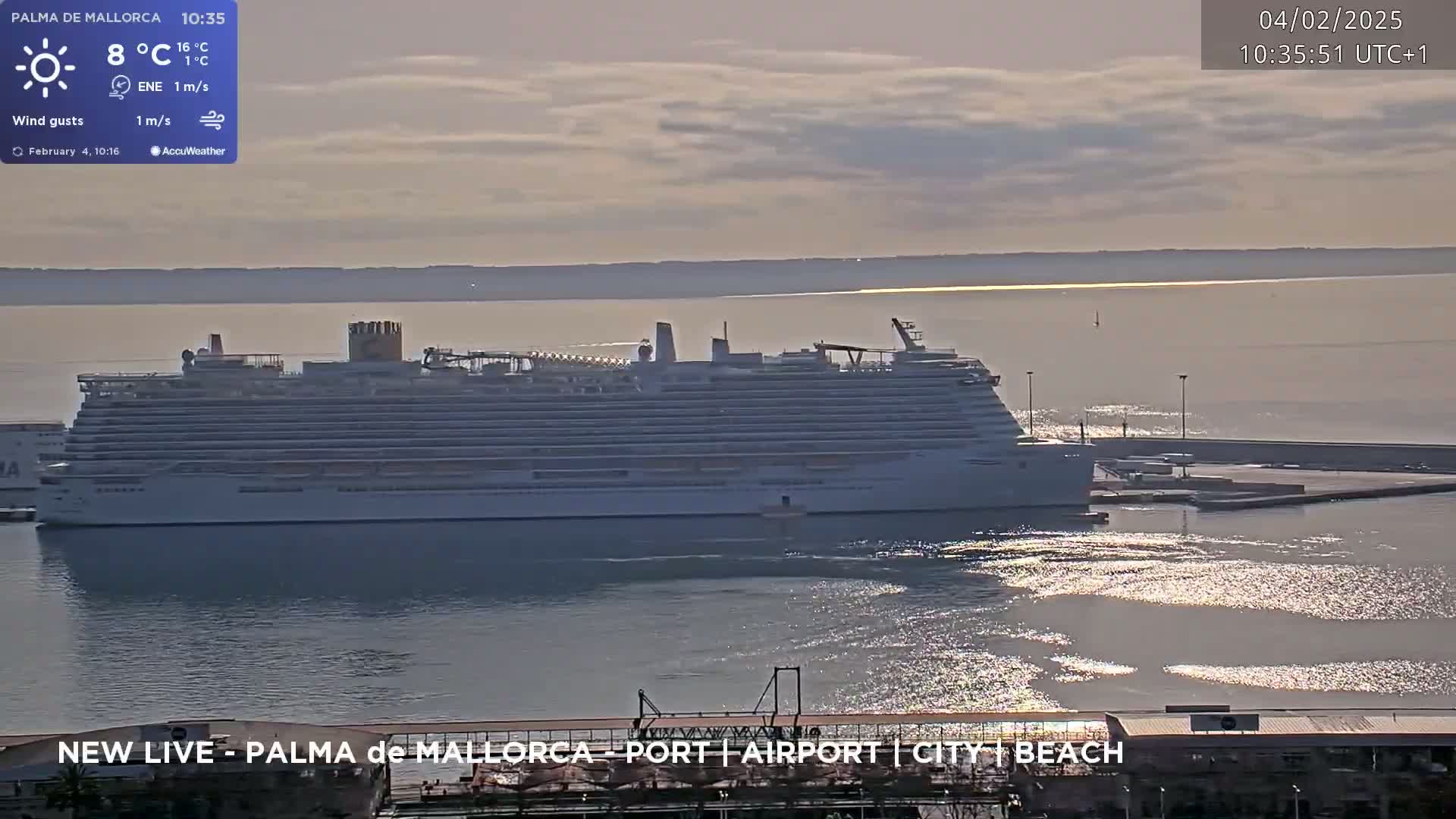 A large cruise ship is docked at a port on a partly sunny day with calm water reflecting sunlight.