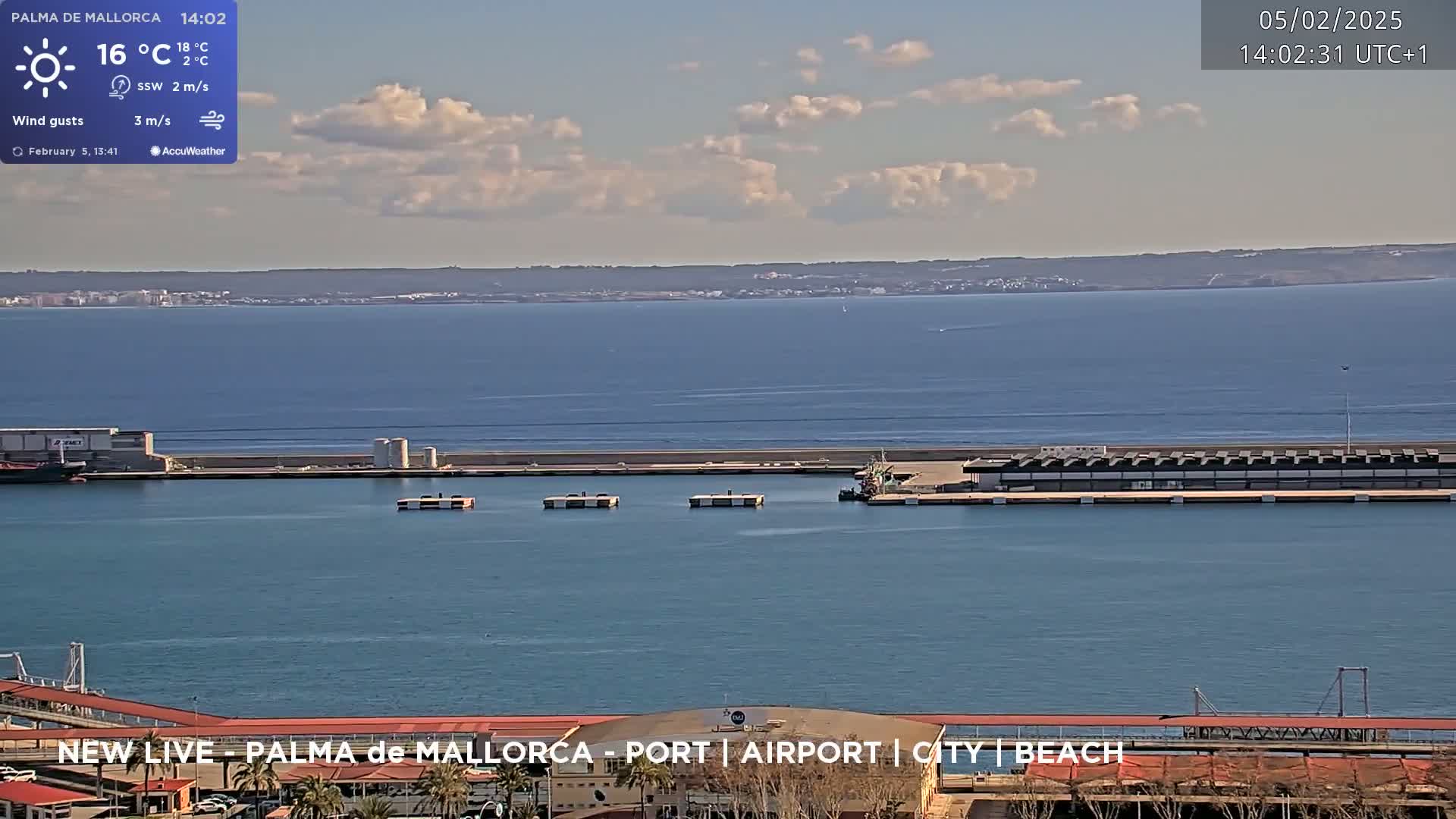 A partly cloudy day shows a harbor with boats and buildings, a calm body of water extending to a distant shoreline.