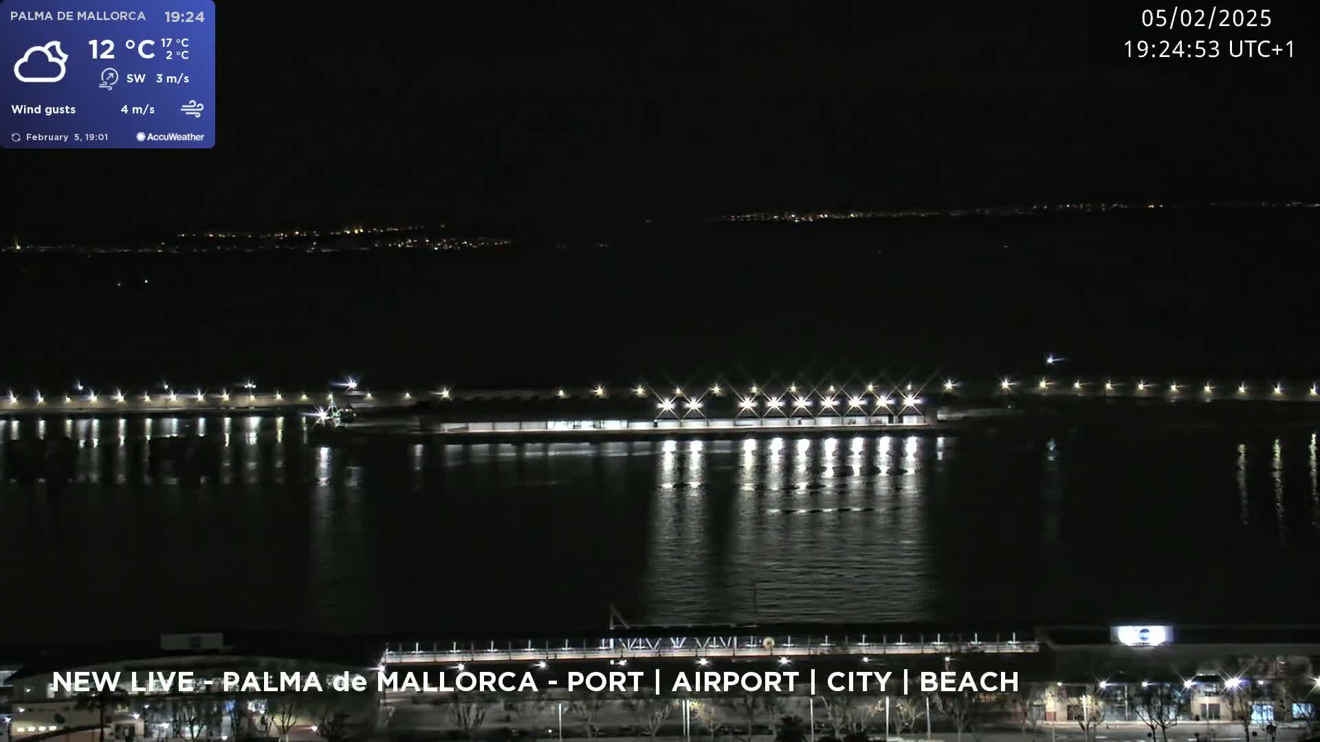 A nighttime view of a calm body of water reflecting the lights of a long pier and distant city lights under a partly cloudy sky.