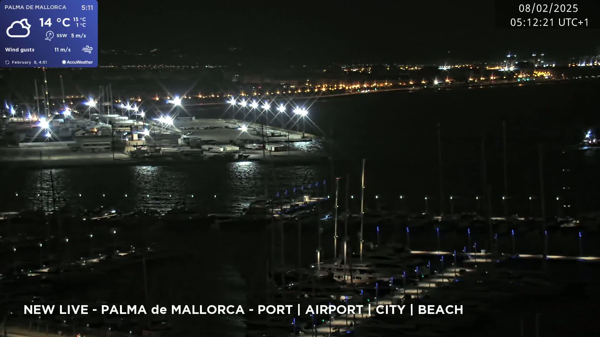 A nighttime view of a harbor filled with boats, illuminated by lights, with a city skyline in the background under partly cloudy skies and a southwest wind.