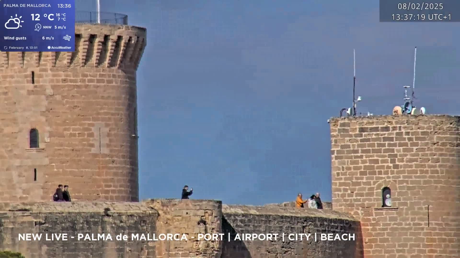 A partly cloudy day reveals stone castle walls and towers with several people standing on top, overlooking a cityscape.
