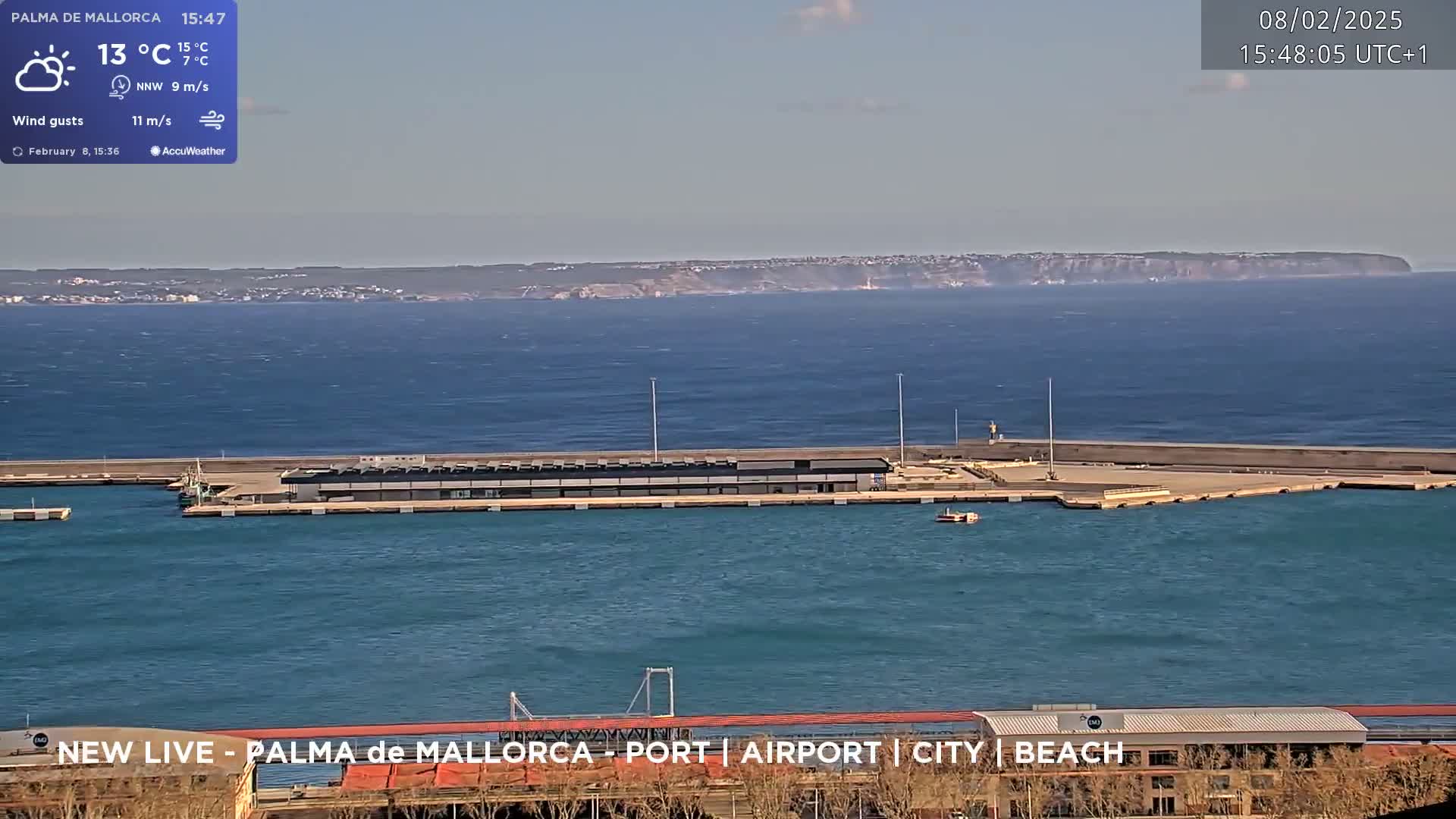 A harbor with a long dock stretches into a calm blue sea, under a partly cloudy sky, with a distant landmass visible on the horizon.