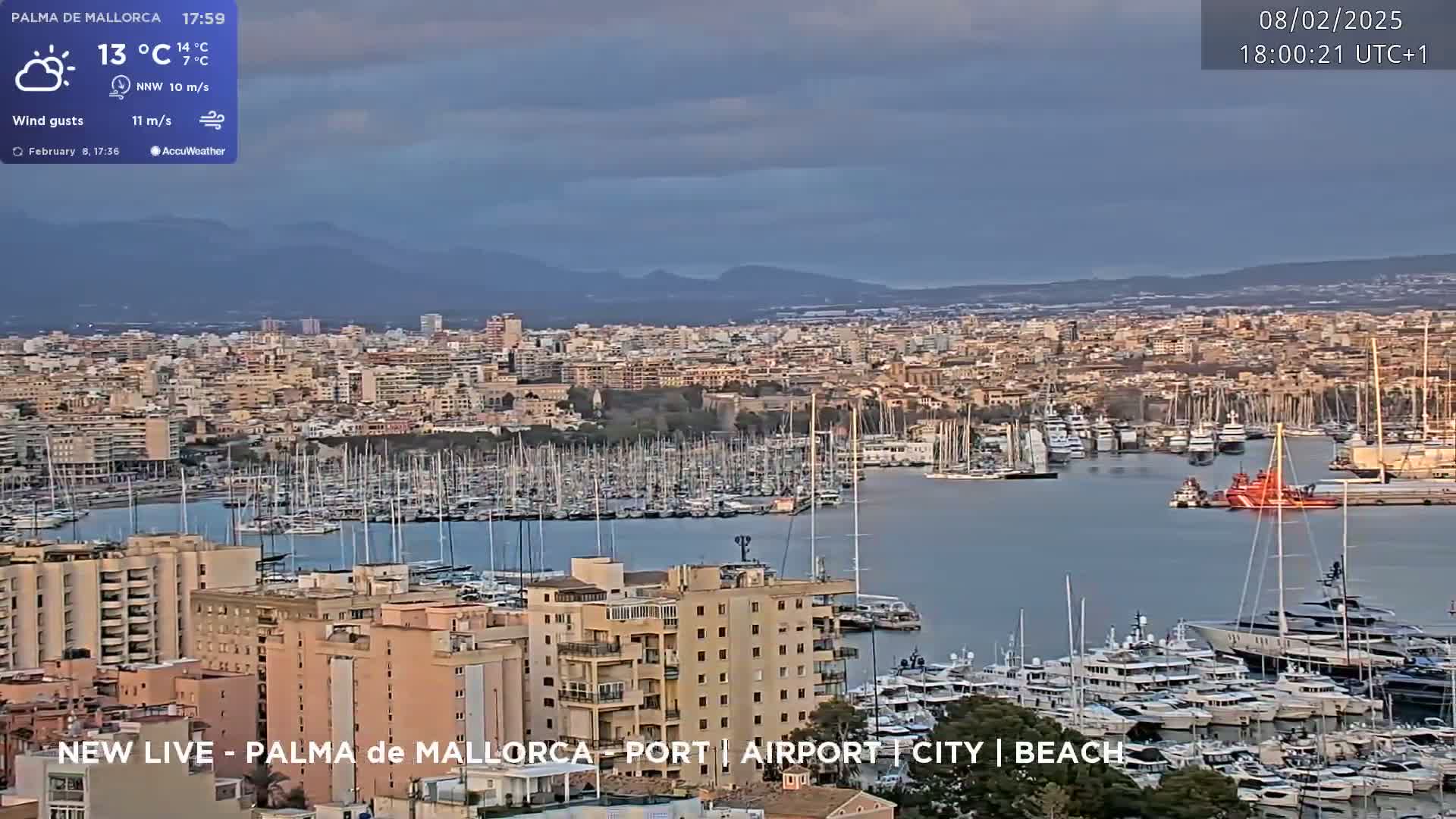 A partly cloudy day overlooks a harbor filled with numerous yachts and boats, next to a city with buildings of varying heights against a backdrop of distant mountains.