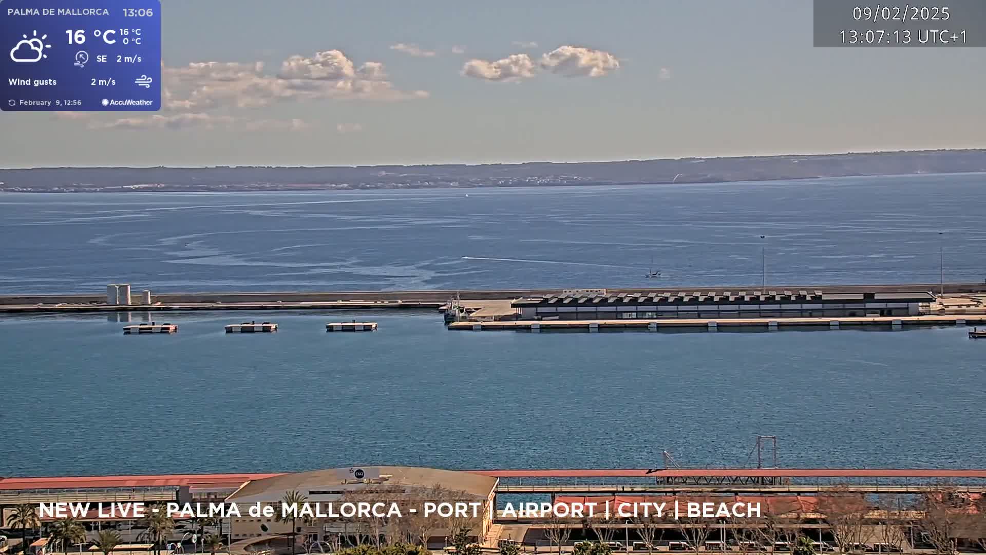 A partly cloudy day shows a harbor with docked boats and buildings, overlooking a calm body of water extending to a distant shoreline.
