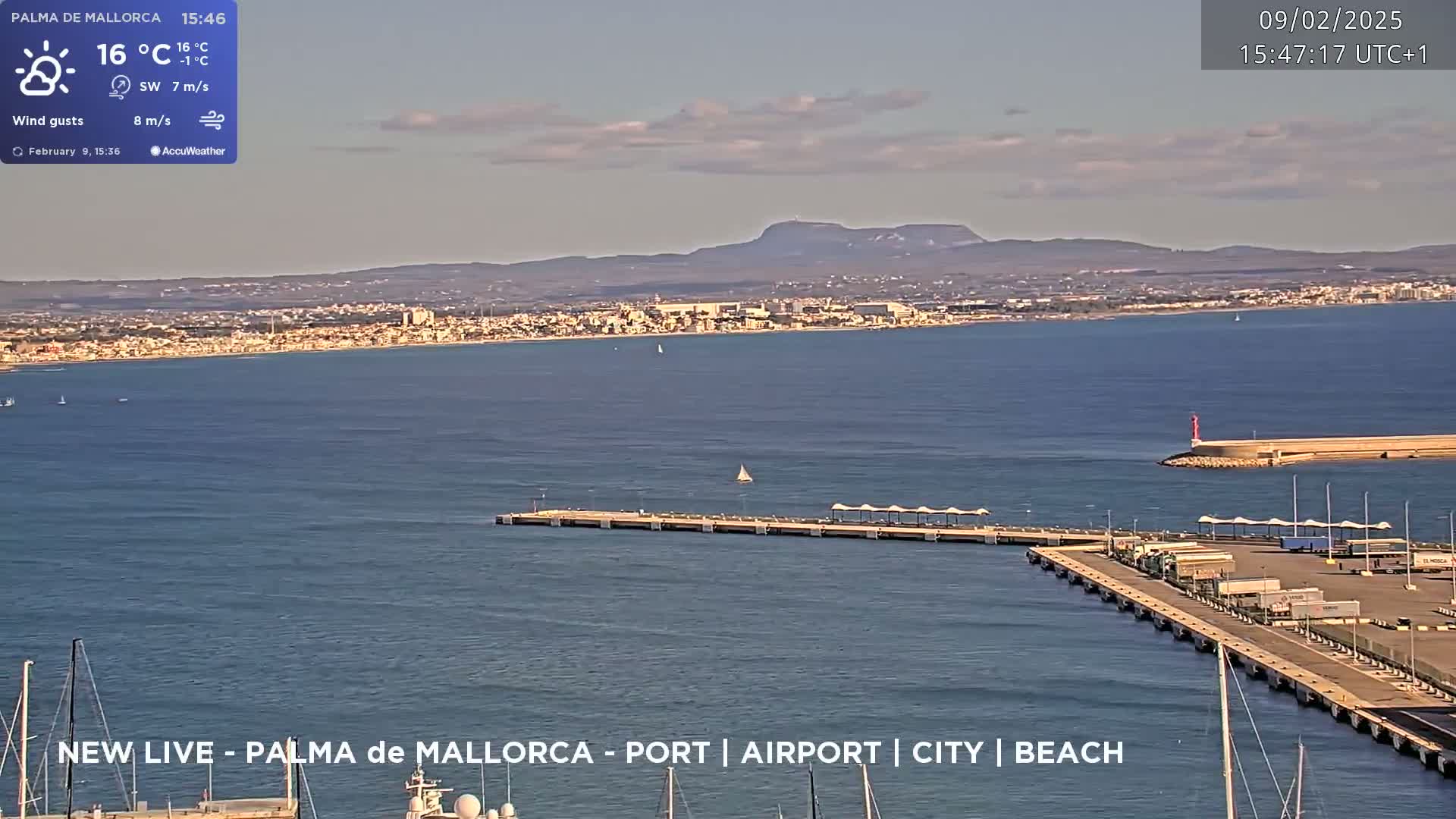 A partly cloudy day shows a coastal city across a calm body of water, with a pier and mountains in the background.