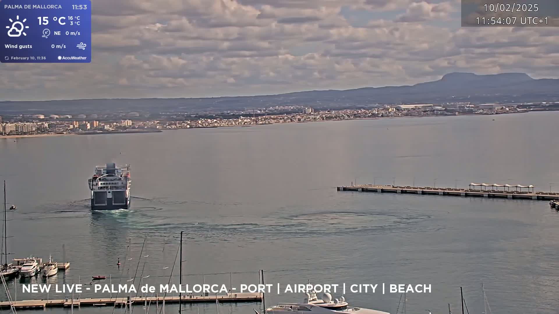 A ferry approaches a harbor in calm waters under a partly cloudy sky, with a city and distant mountains visible in the background.