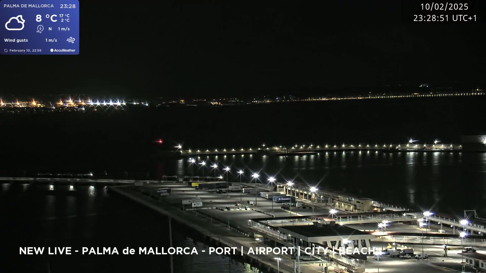 A nighttime aerial view of a port area with calm water, illuminated by numerous lights, and a city skyline in the distance.