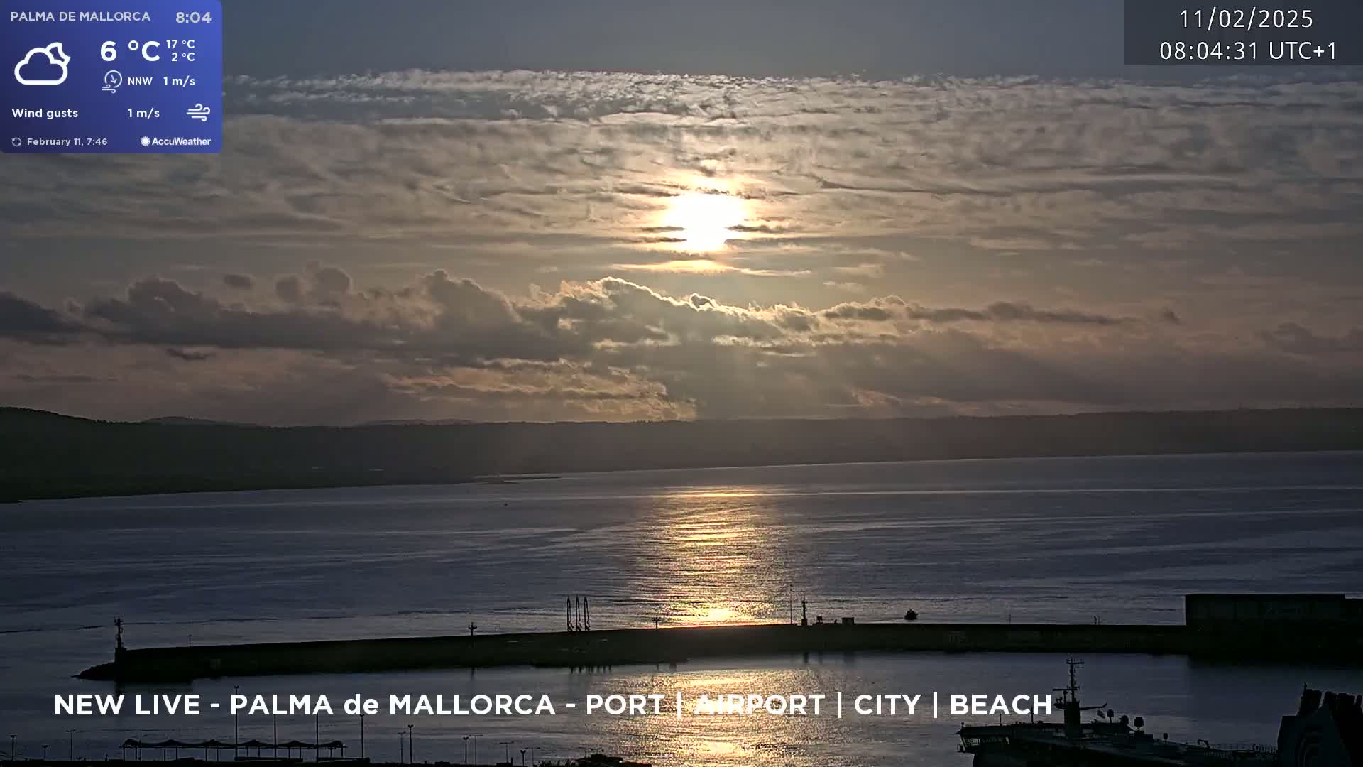 A partly cloudy sunrise casts a golden glow on a calm body of water, reflecting the light near a harbor.