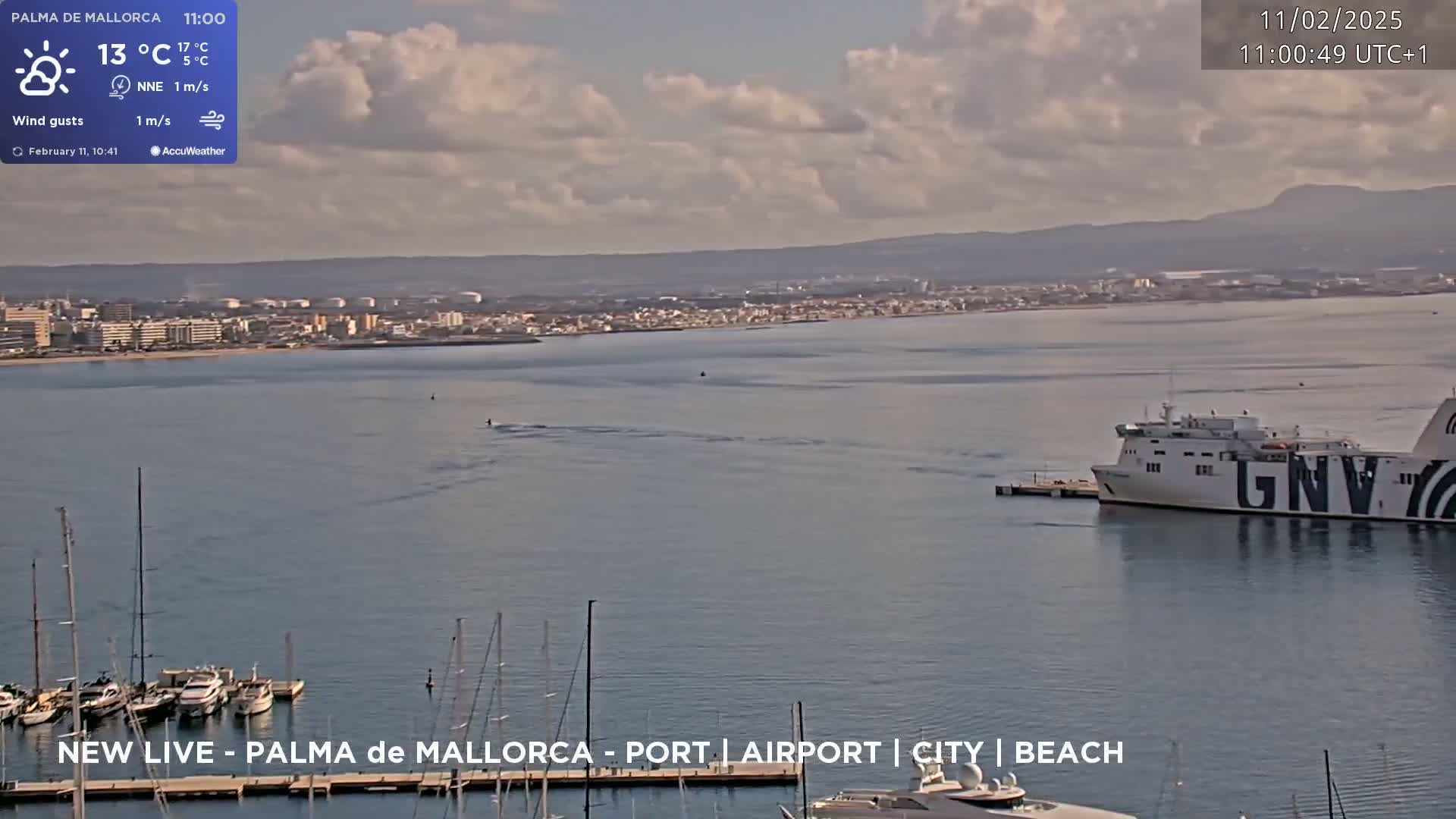 A partly cloudy day shows a large ferry docked near a harbor filled with sailboats and yachts, with a city and distant mountains visible in the background.