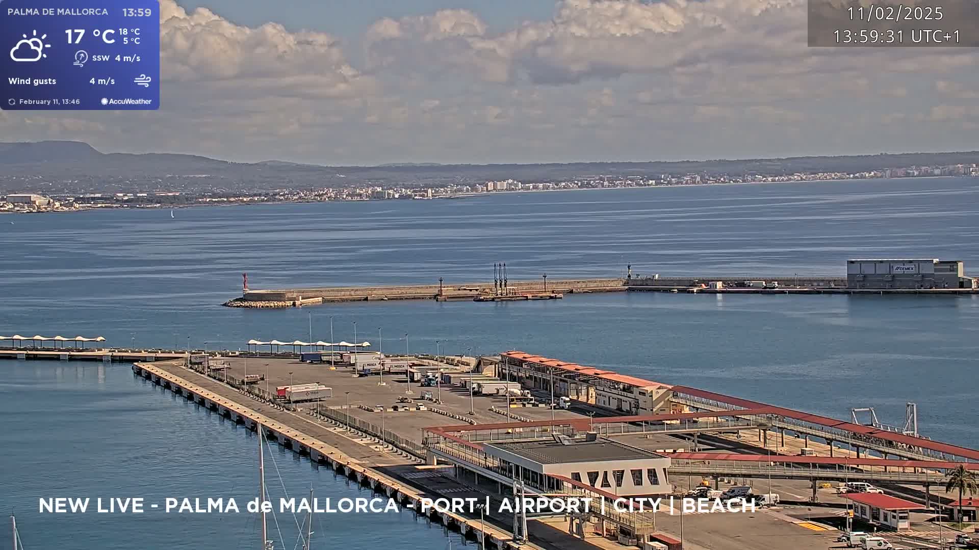 A partly cloudy day shows a port with numerous vehicles and buildings, overlooking a calm body of water and a distant city under a mostly cloudy sky.