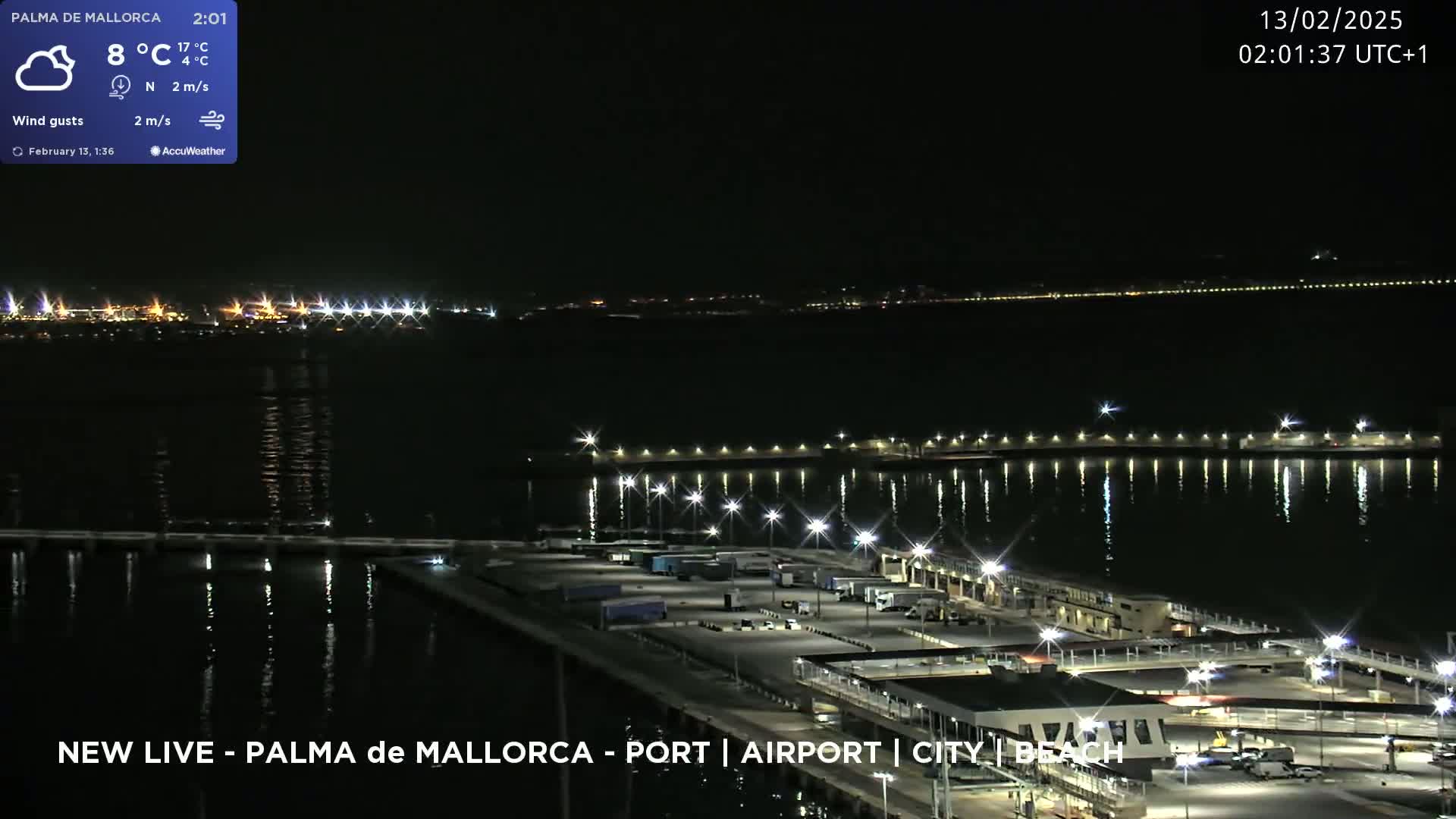 A nighttime, aerial view of a port with calm waters reflecting lights, under a cloudy sky with light wind gusts.