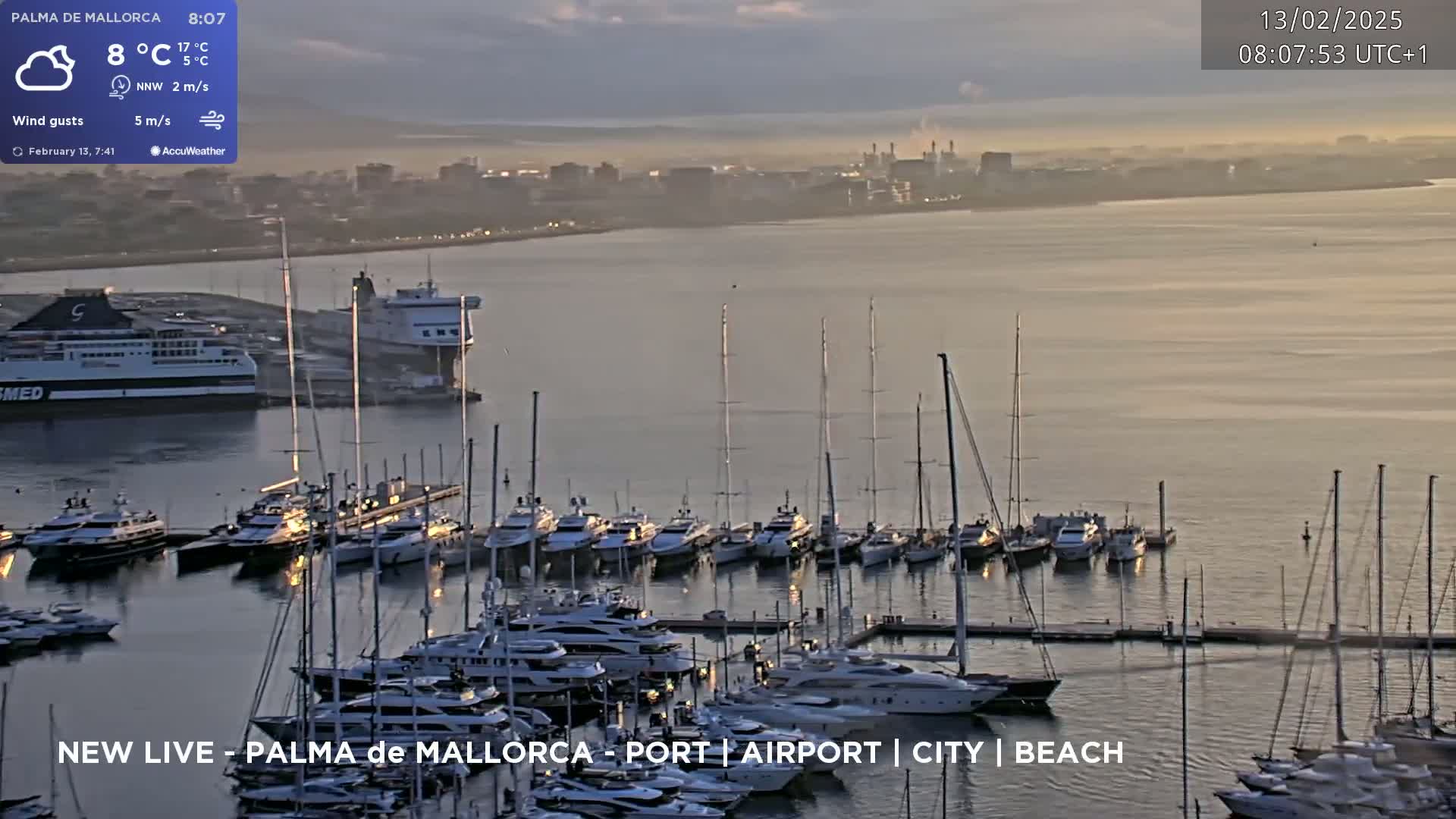 A harbor filled with many yachts and sailboats is shown at sunrise with a hazy city in the distance under partly cloudy skies.