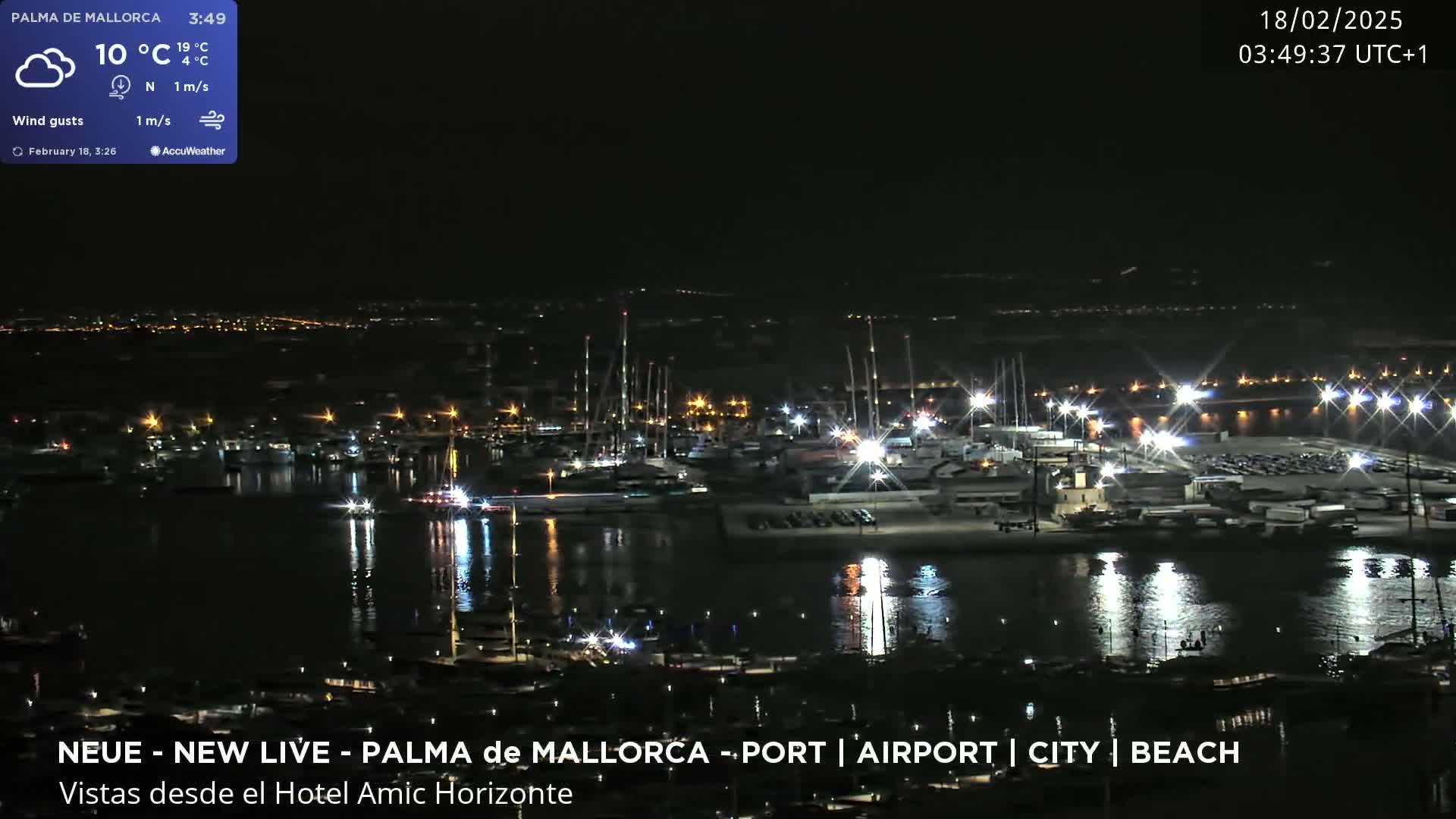 A nighttime view of a harbor filled with boats and lit by many lights, with a city in the background under partly cloudy skies.