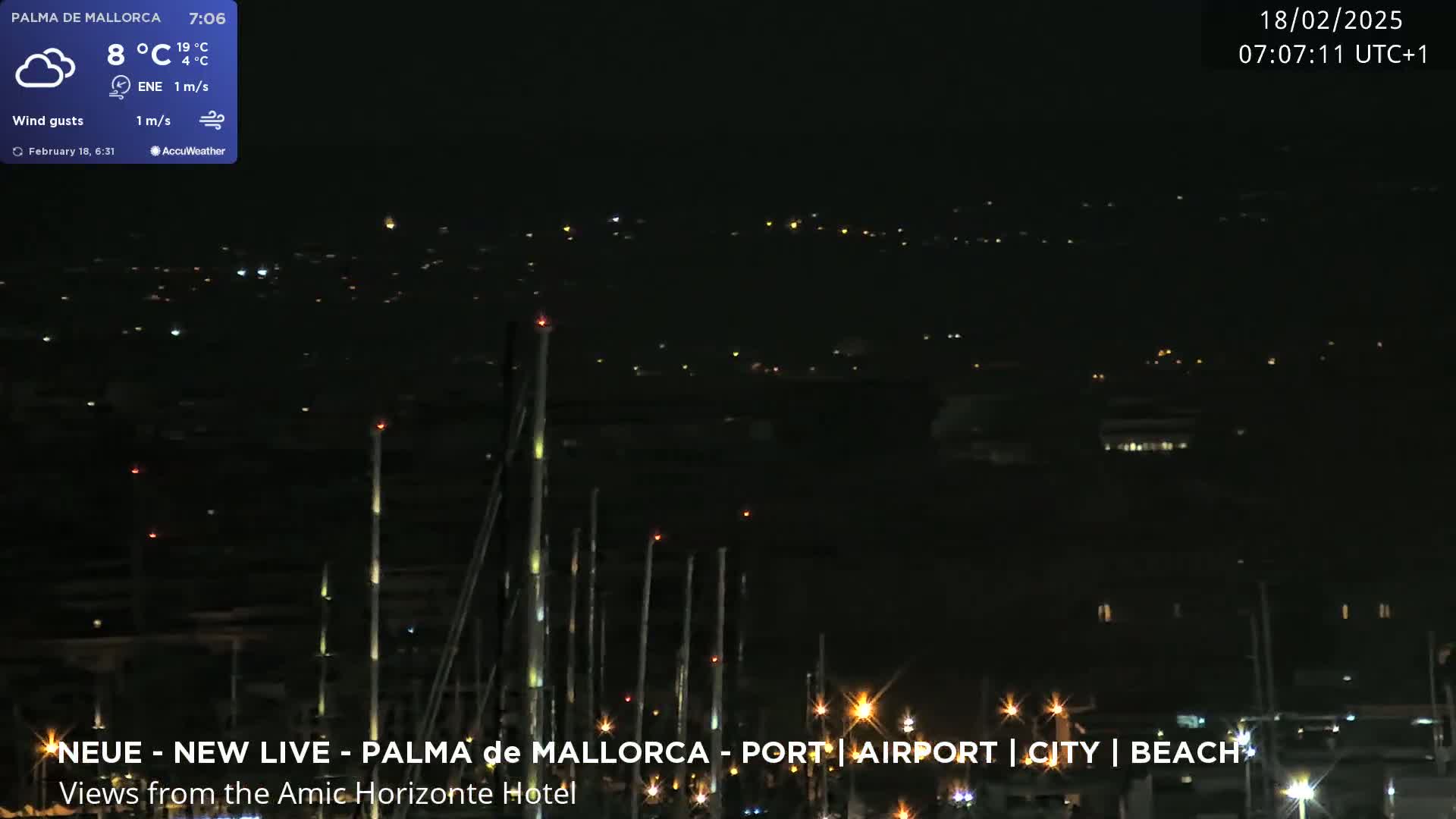 A nighttime view of a harbor with many masts, sparsely lit buildings in the distance, and a partly cloudy sky.