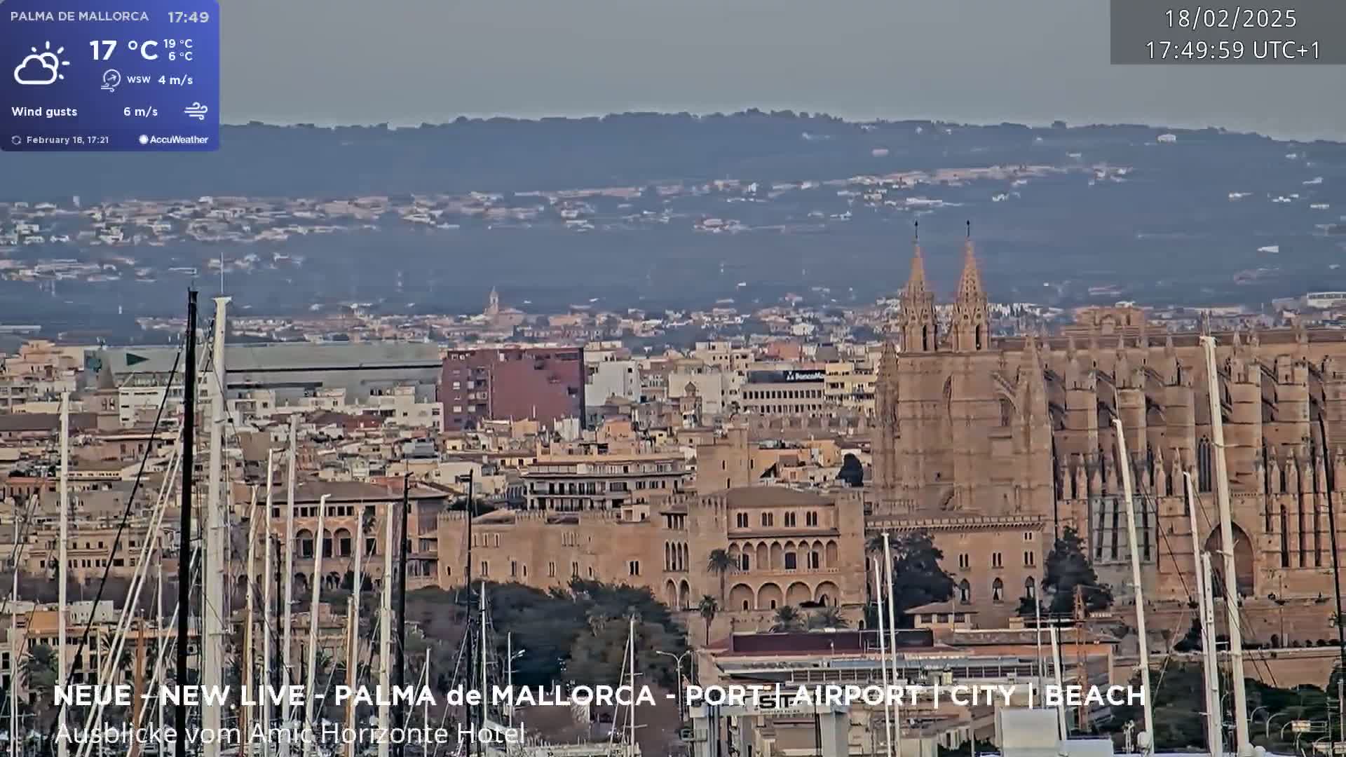 A partly cloudy day reveals a harbor filled with sailboats, a city skyline, and a large stone cathedral, all set against a hazy mountain backdrop.