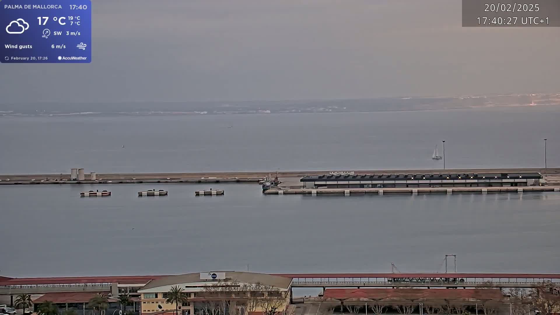 A harbor scene on a cloudy day shows a calm body of water with a long dock, several small boats, and a distant shoreline.