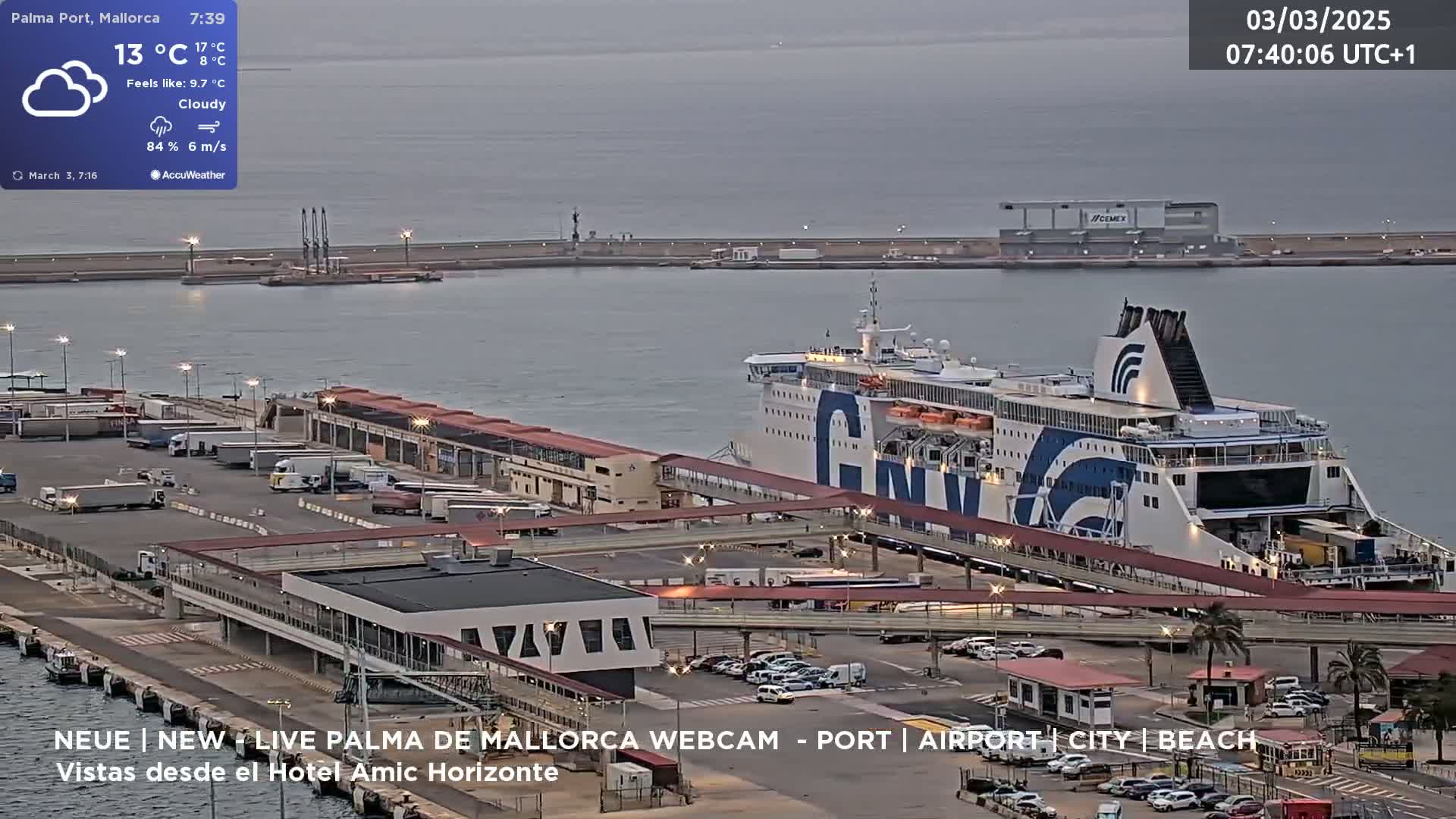A large ferry is docked at a port under cloudy skies, with numerous vehicles parked nearby.