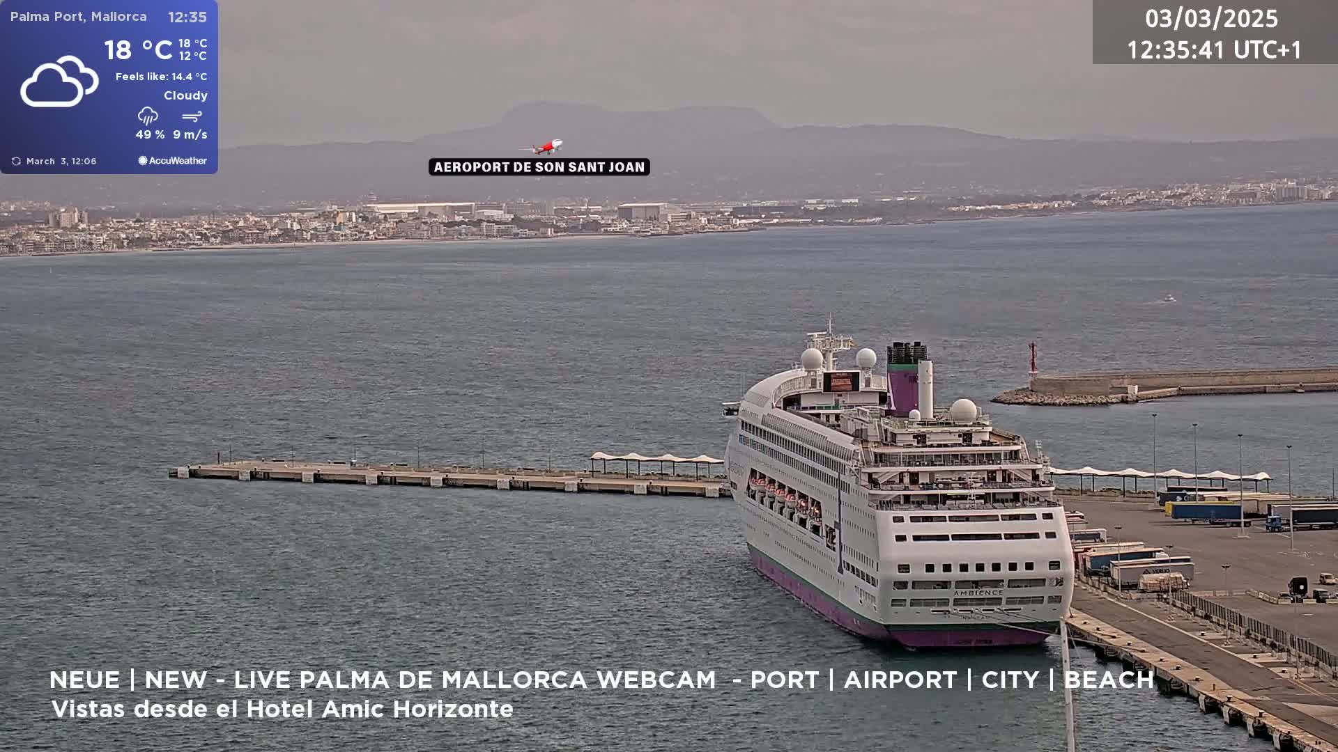 A large cruise ship is docked at a port on a cloudy day, with a city and distant mountains visible in the background.