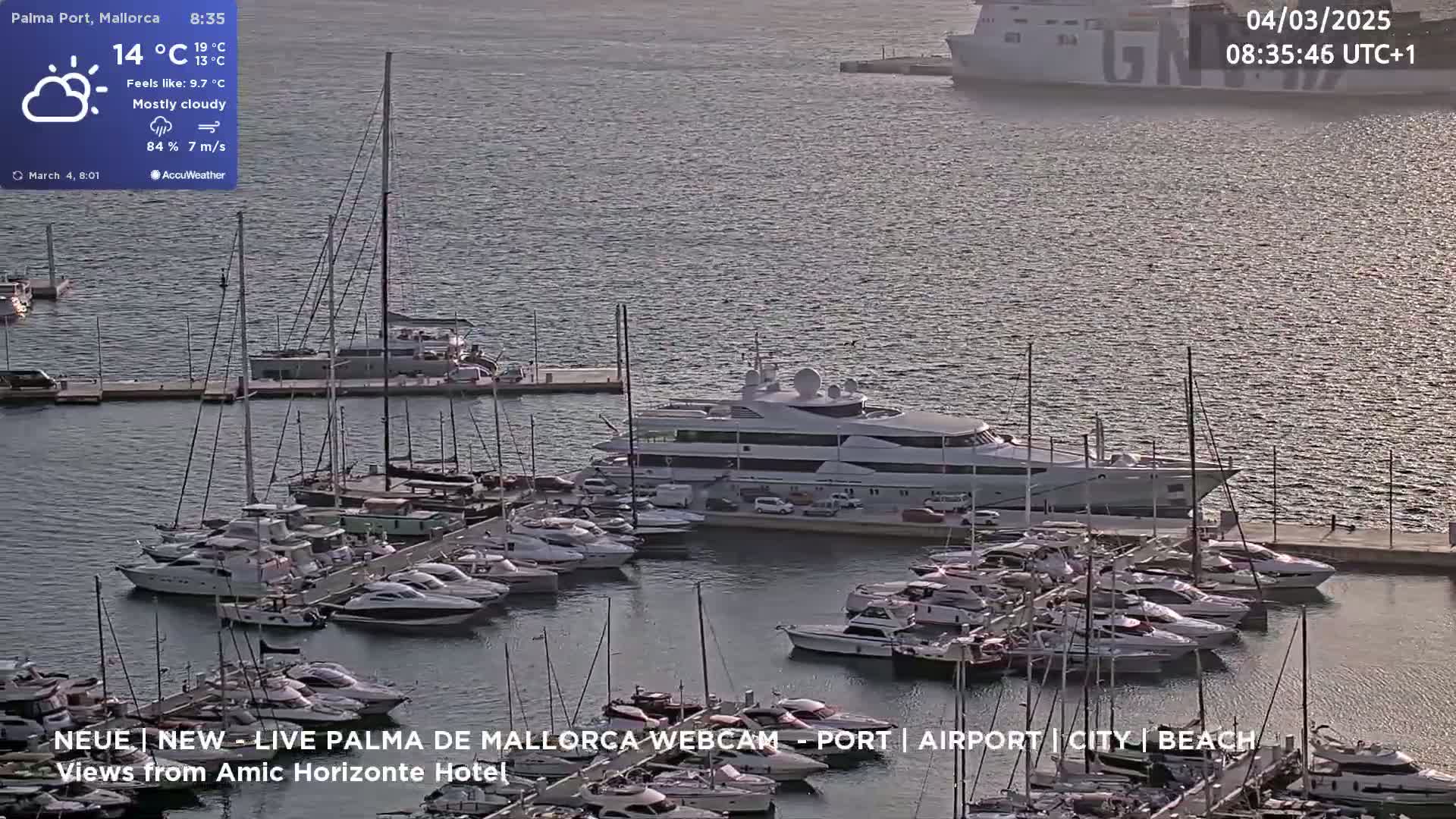A mostly cloudy harbor scene shows many yachts and boats docked, with a large yacht prominently featured in the center, and a large ship in the background.