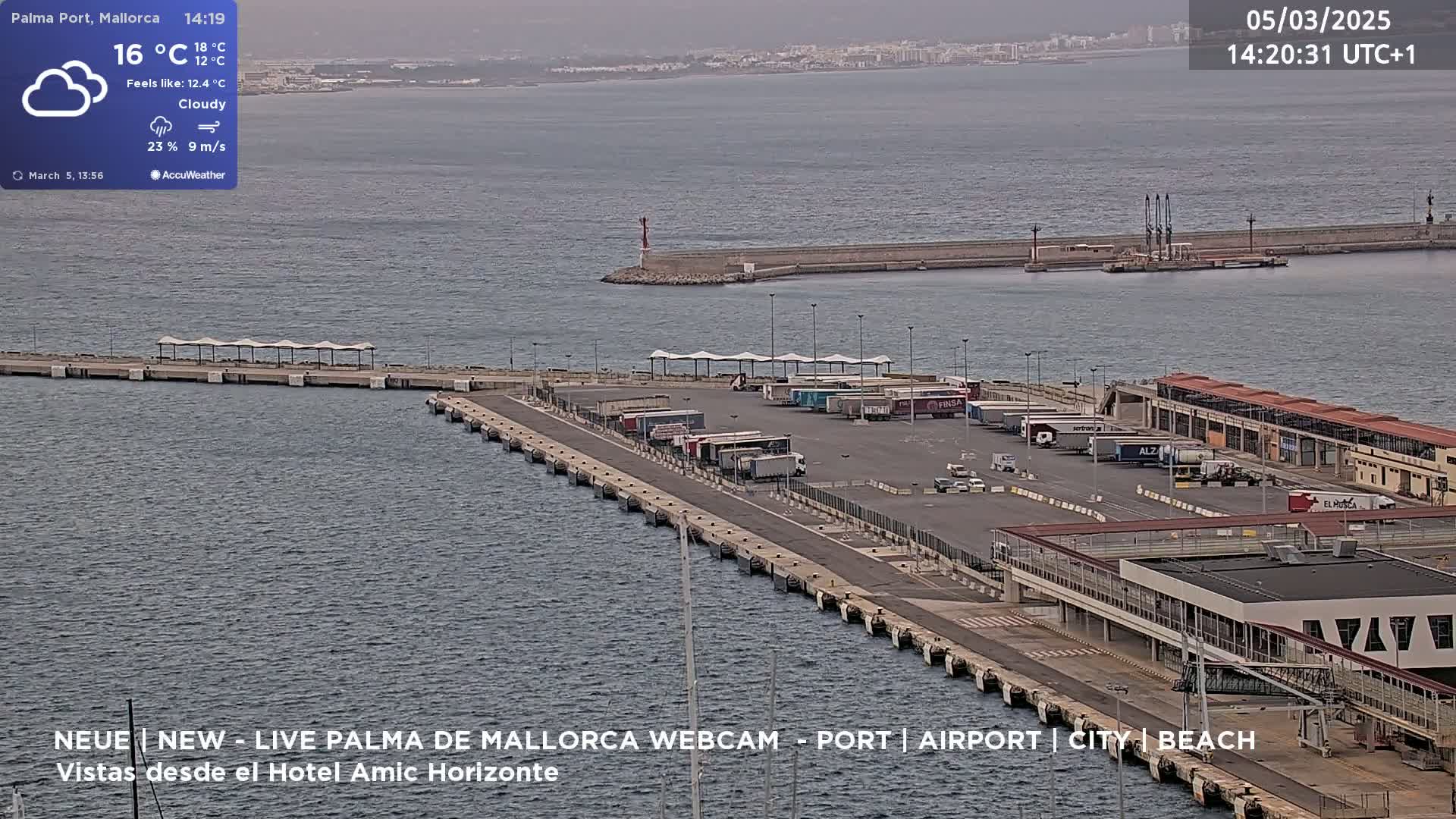 An overcast day shows a port with numerous trucks parked near buildings along a long pier extending into a body of water.