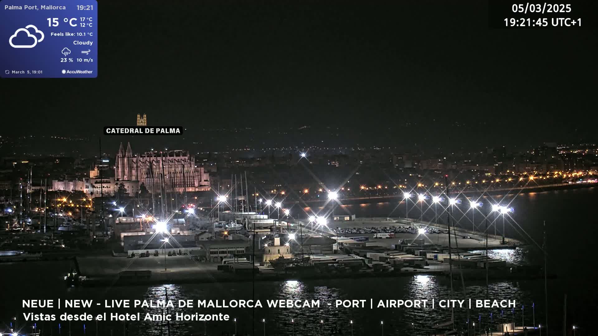 A nighttime, cloudy view of a city harbor with brightly lit boats and buildings, including a large cathedral, under partly cloudy skies.