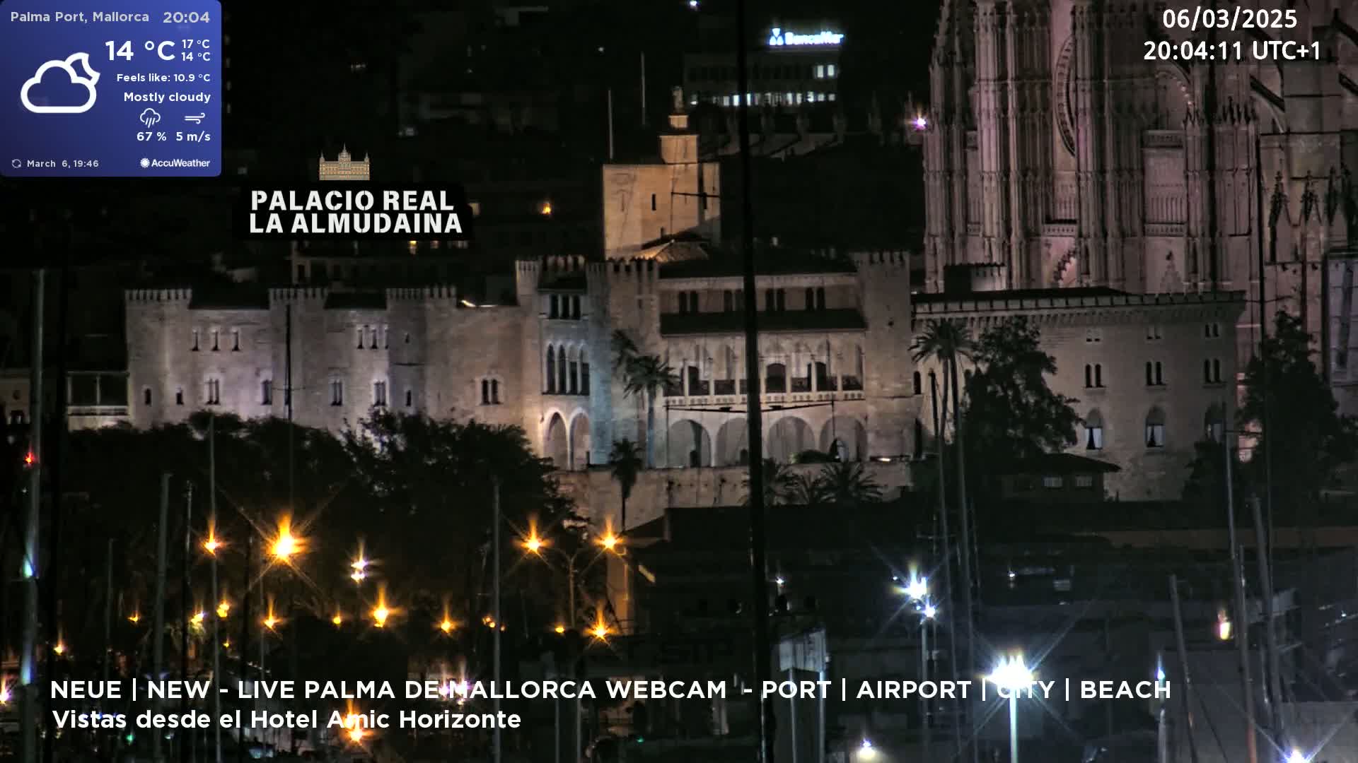 A nighttime long shot of a city harbor, featuring a large illuminated stone building and a cathedral under mostly cloudy skies.