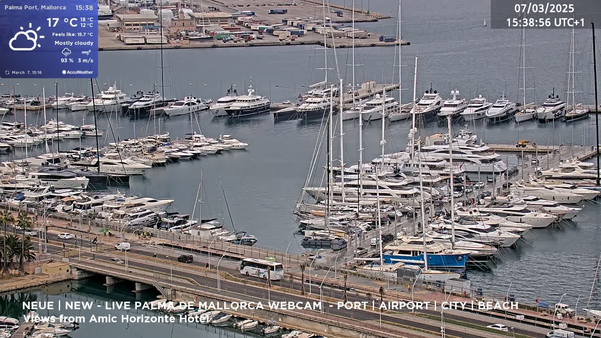 A mostly cloudy day shows a marina filled with many yachts and sailboats docked, with a roadway and a bus visible in the foreground.