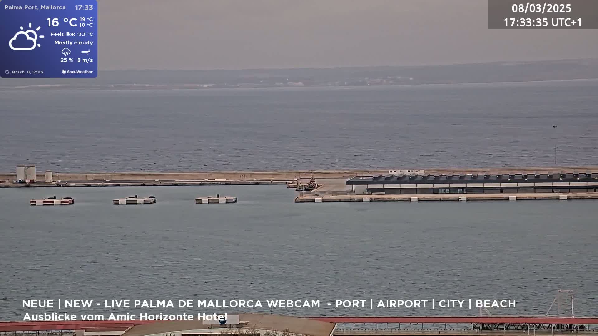 A mostly cloudy day shows a harbor with several docked boats and a large building along the waterfront.