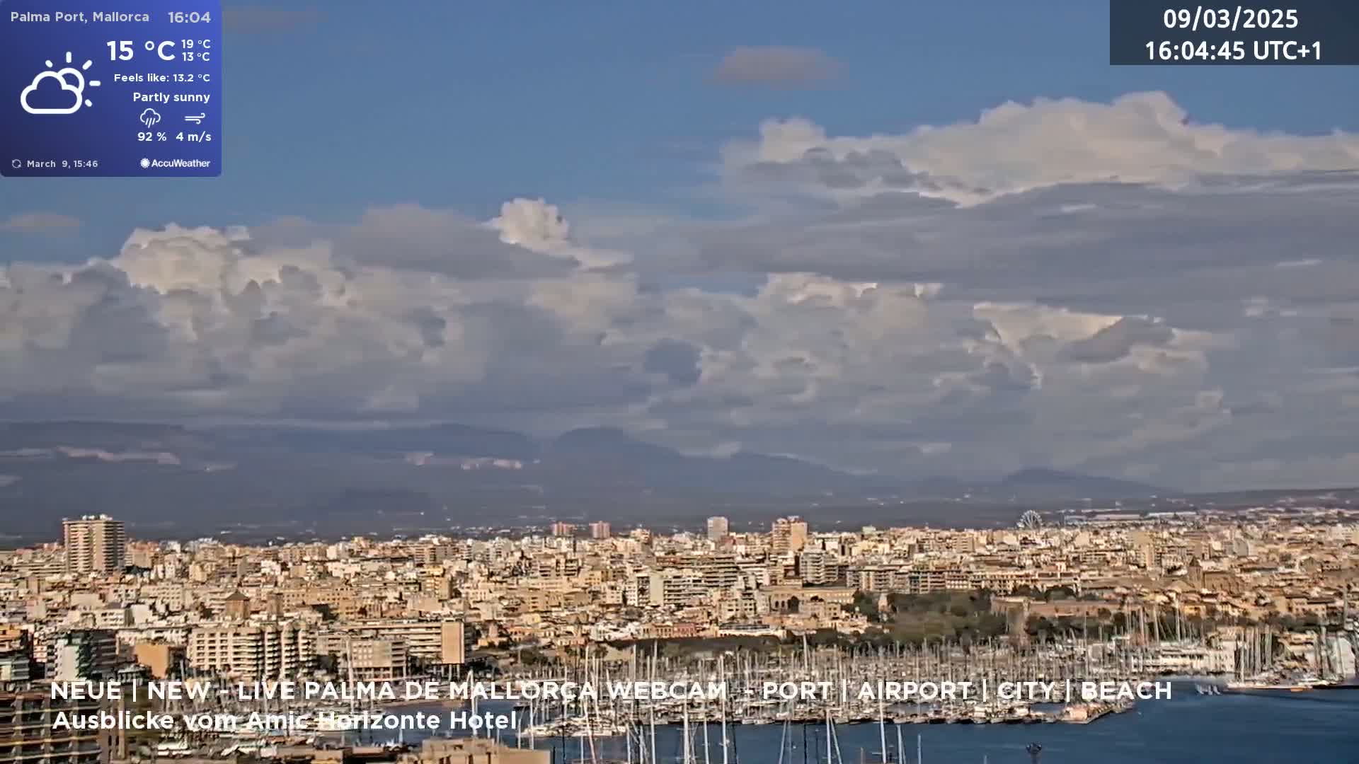 A partly sunny, windy day reveals a panoramic view of a city harbor filled with boats, backed by buildings and distant mountains.