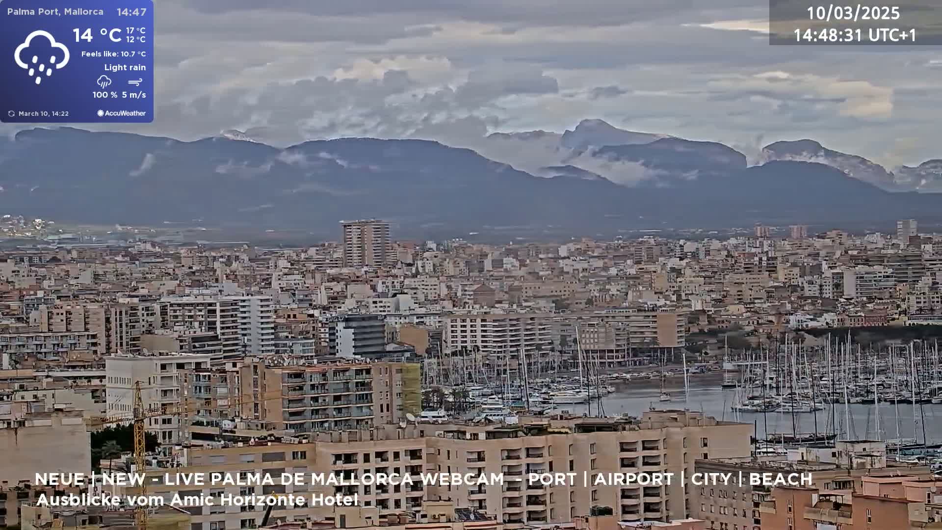 A city harbor filled with sailboats is seen below a mountain range partially obscured by light rain and clouds.