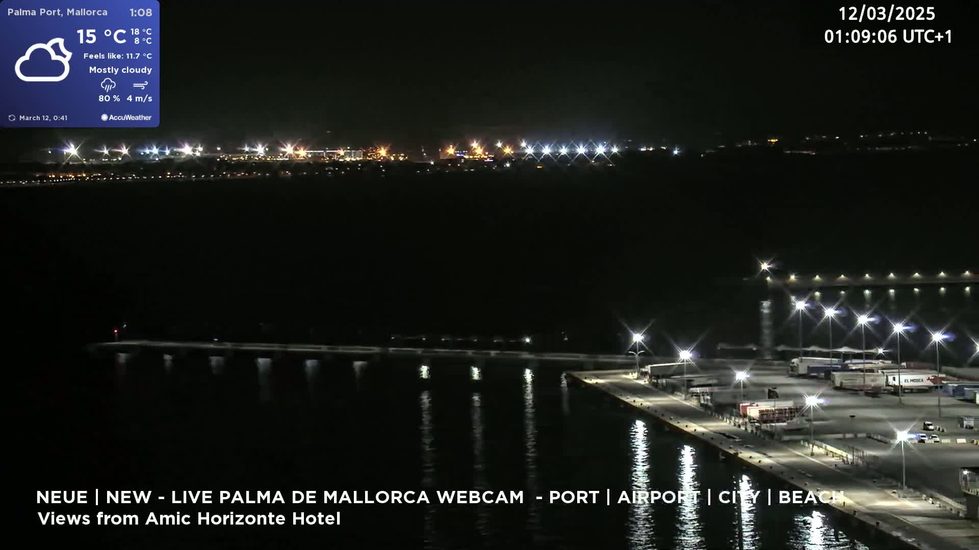 A nighttime view of a mostly cloudy port area with calm water reflecting the lights of the dock and distant city.