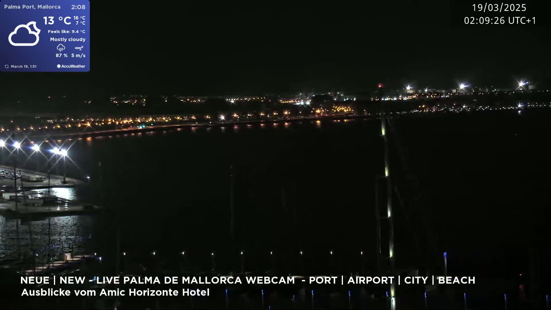 A nighttime, mostly cloudy view of a city and harbor illuminated by lights, with a calm body of water in the foreground.