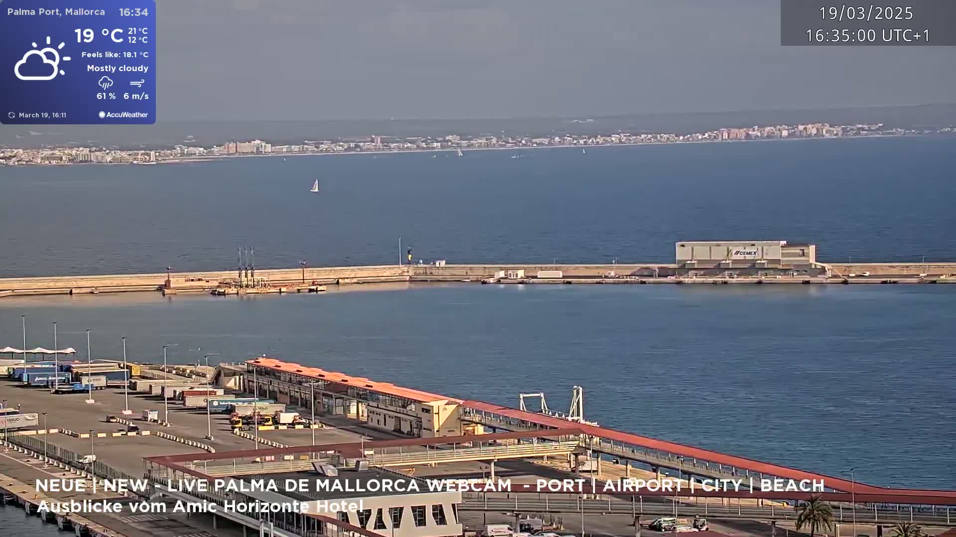 A mostly cloudy day shows a port with a long pier extending into a calm body of water, with a distant city skyline visible.