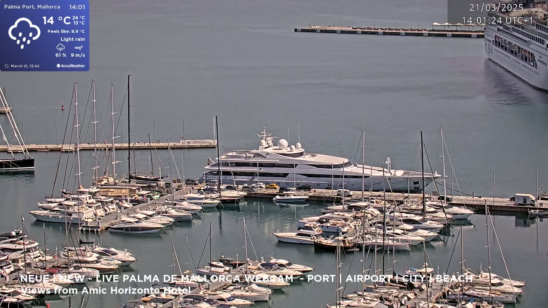 A marina filled with numerous yachts and sailboats under light rain, with a large luxury yacht prominently docked and a cruise ship visible in the distance.