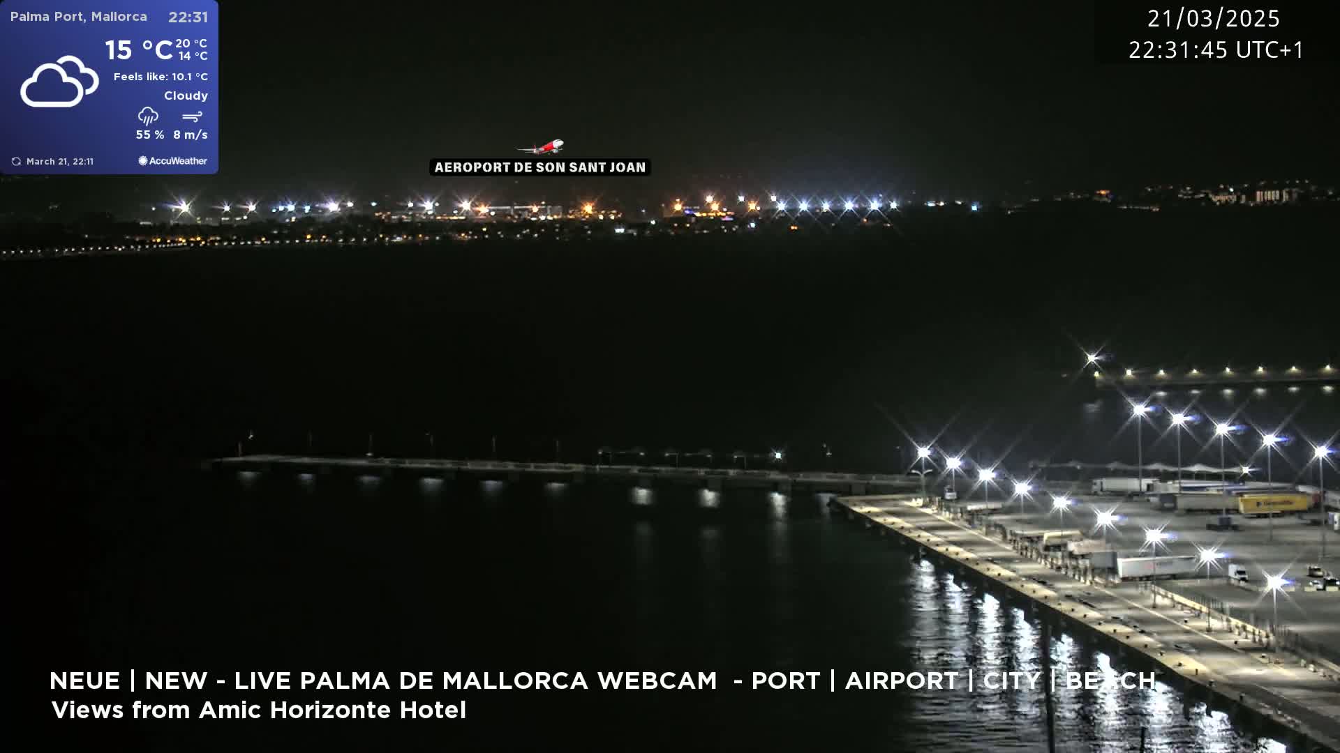 A nighttime view of a port with brightly lit docks and a city skyline in the distance under cloudy skies.
