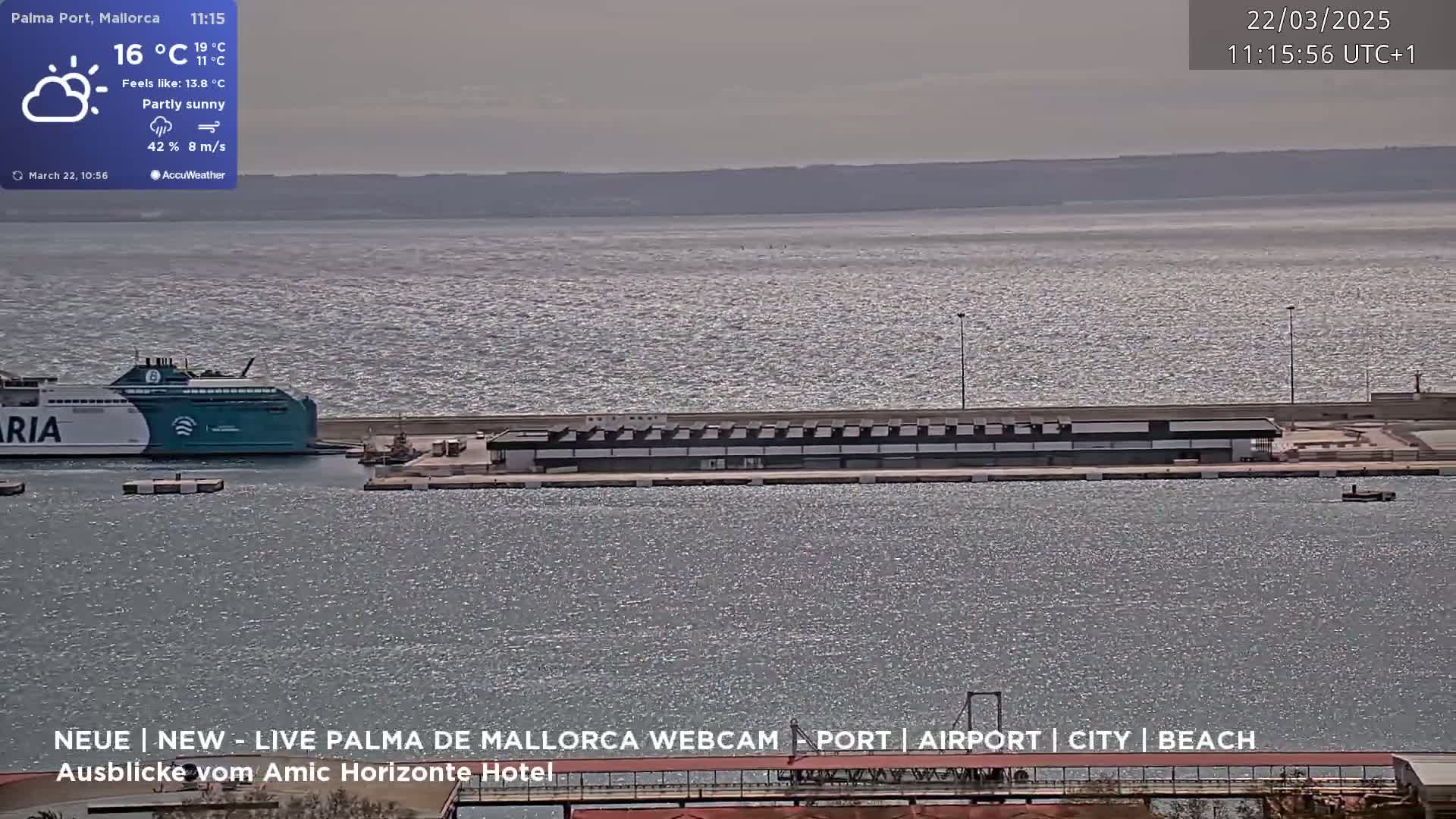 A partly sunny day shows a teal ferry docked at a port with a long building along the water's edge.