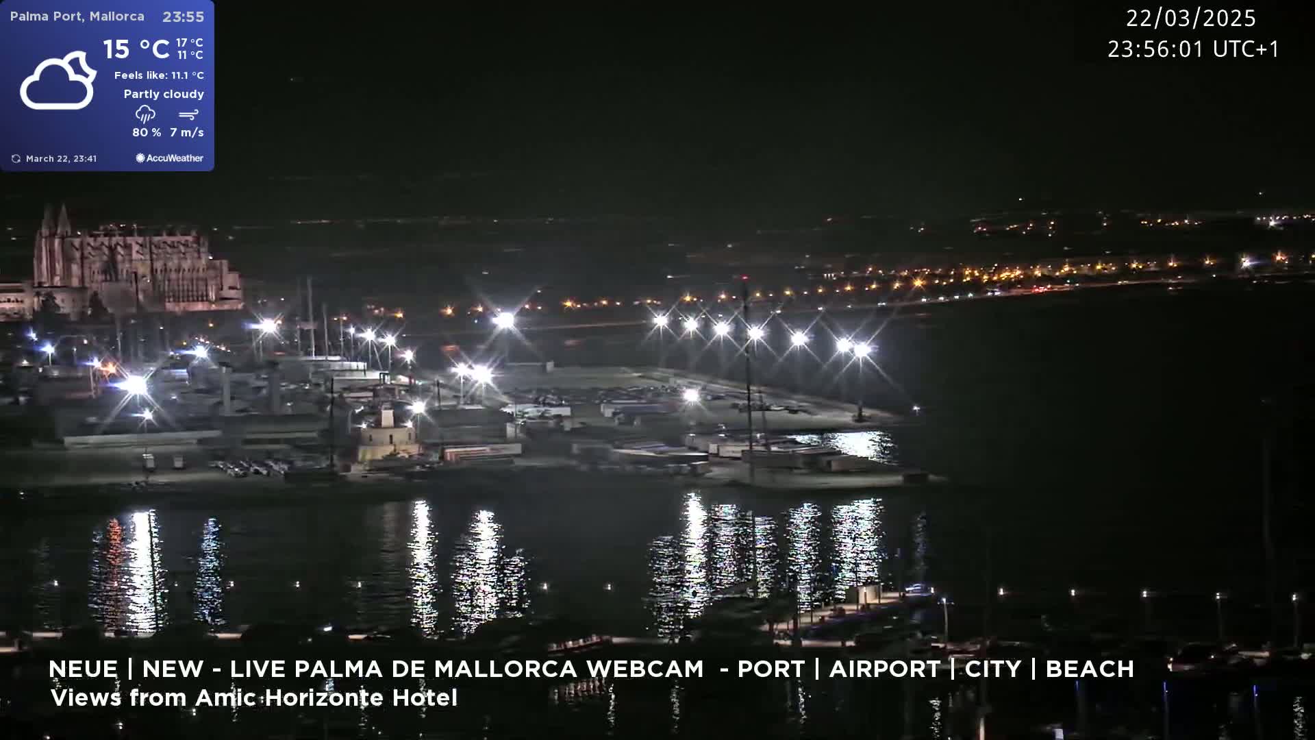A nighttime, partly cloudy view of a harbor with brightly lit boats and a large building in the background, reflecting light on calm water.