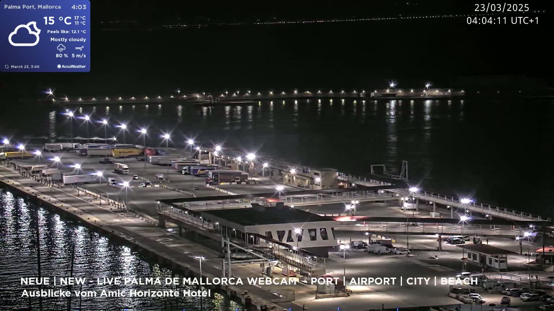 A nighttime, mostly cloudy view of a port with many vehicles parked, illuminated by numerous lights, next to dark water reflecting the lights.