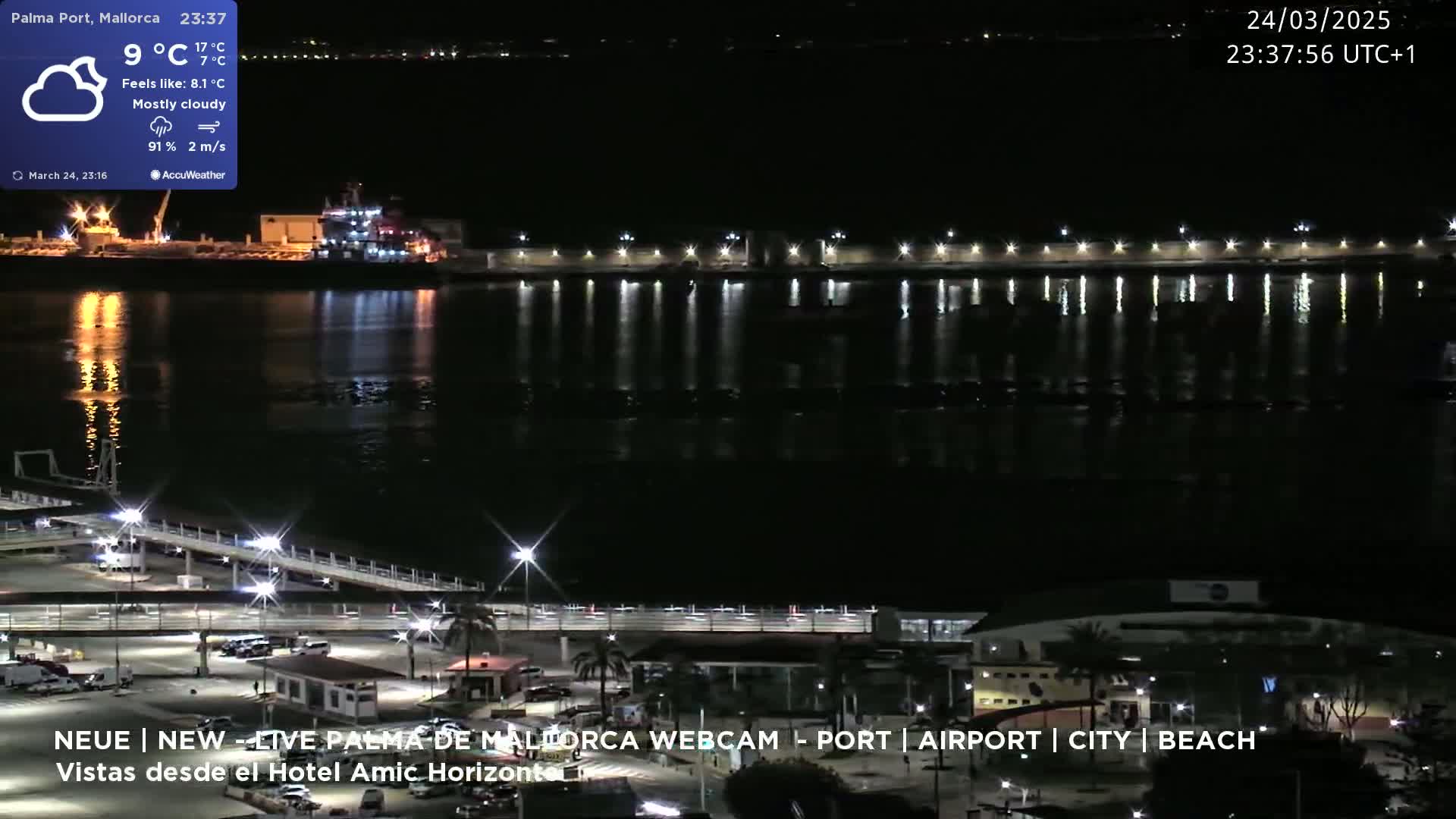 A nighttime view of a harbor with a mostly cloudy sky, showing a body of water reflecting lights from the shore and buildings.