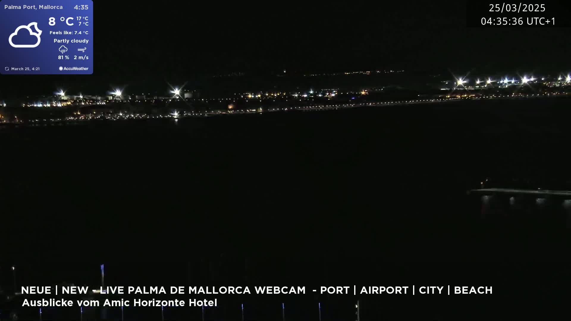 A nighttime view of a city's lights along a dark body of water under partly cloudy skies.