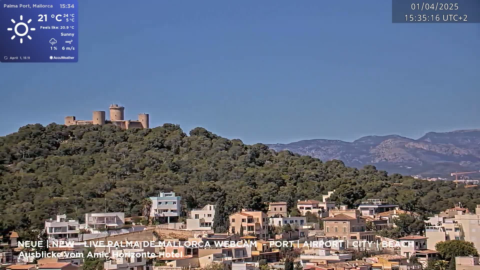A sunny day reveals a coastal town nestled below a hilltop castle, with mountains in the background.