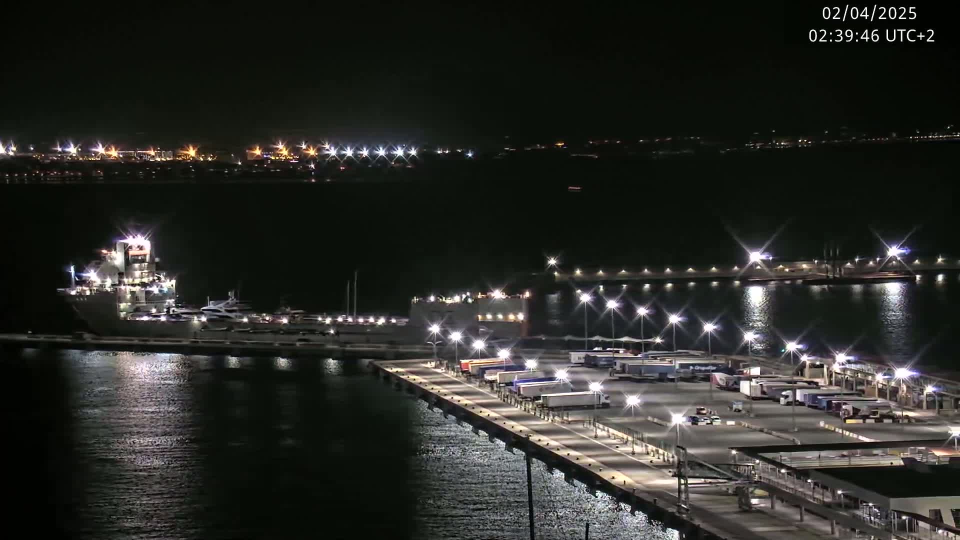 A large ship is docked at a brightly lit port at night, with numerous semi-trucks parked nearby.