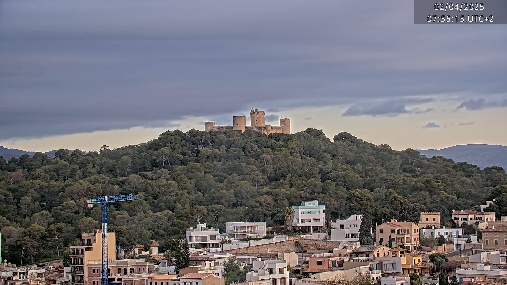 A stone castle sits atop a tree-covered hill overlooking a town under a cloudy sky.