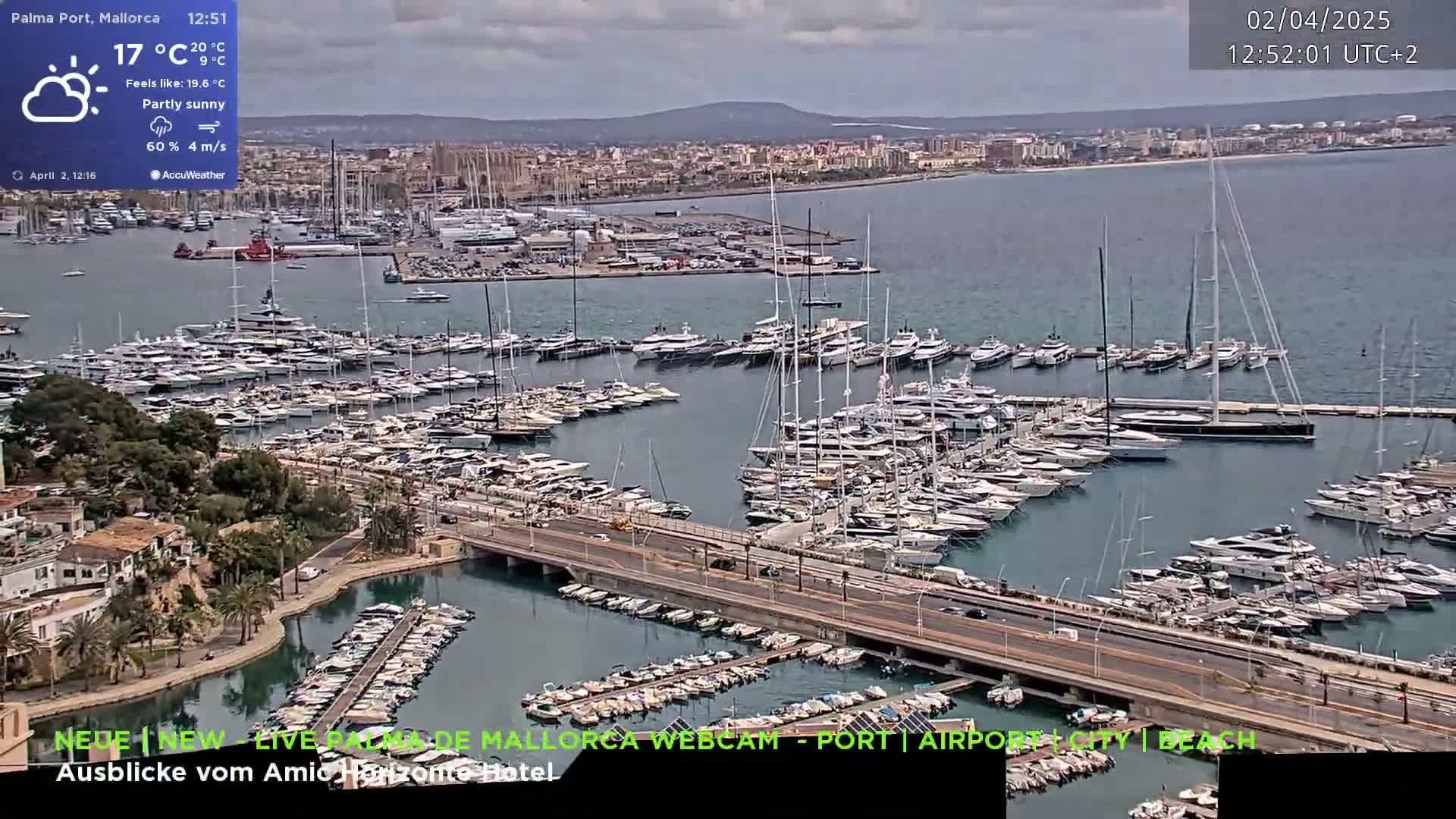 A partly sunny day shows a harbor filled with numerous yachts and sailboats, a city in the background, and a bridge connecting two areas of the marina.
