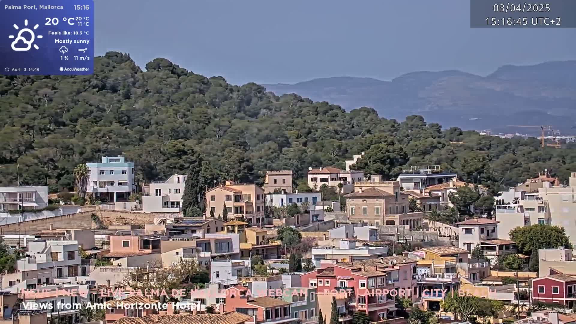 A mostly sunny, breezy day overlooks a hillside town with numerous houses clustered together, backed by a verdant hill and distant mountains.