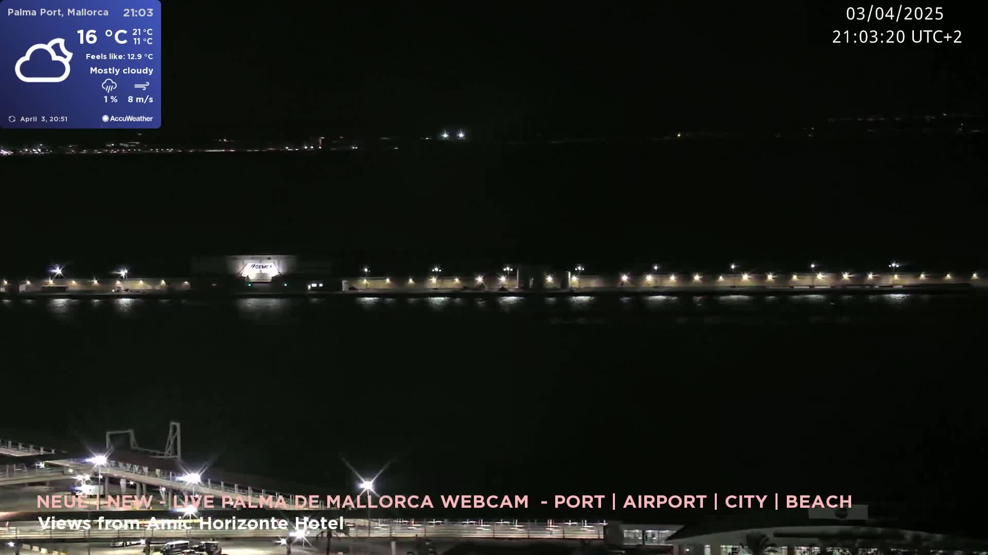 A mostly cloudy nighttime scene shows a dark body of water with a brightly lit waterfront area and distant lights on the other side.